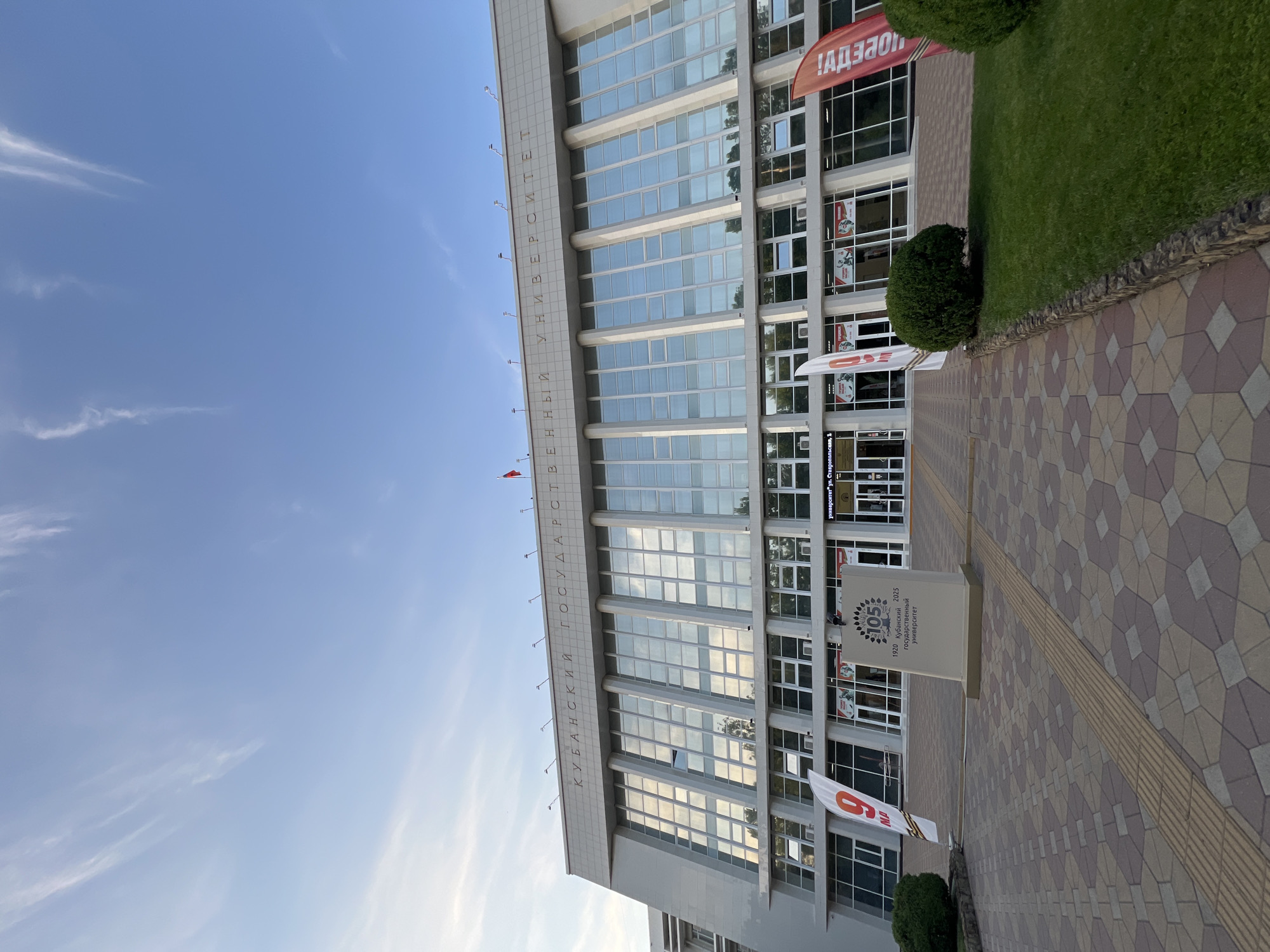 The image shows the front facade of the Kuban State University building in Krasnodar, Russia. The building is a modern, multi-story structure with a large glass facade. The inscription on the top of the building reads "КУБАНСКИЙ ГОСУДАРСТВЕННЫЙ УНИВЕРСИТЕТ" (Kuban State University). In front of the building, there is a paved walkway with a tactile paving strip. Several flags are visible, including one with the number "9" and "мая" (May 9th), and another red flag with text. A stone monument with "105" and "1920-2025 Государственный университет" (1920-2025 State University) is also present. There are also manicured bushes and a lawn in front of the building. The sky is clear blue with some wispy clouds.