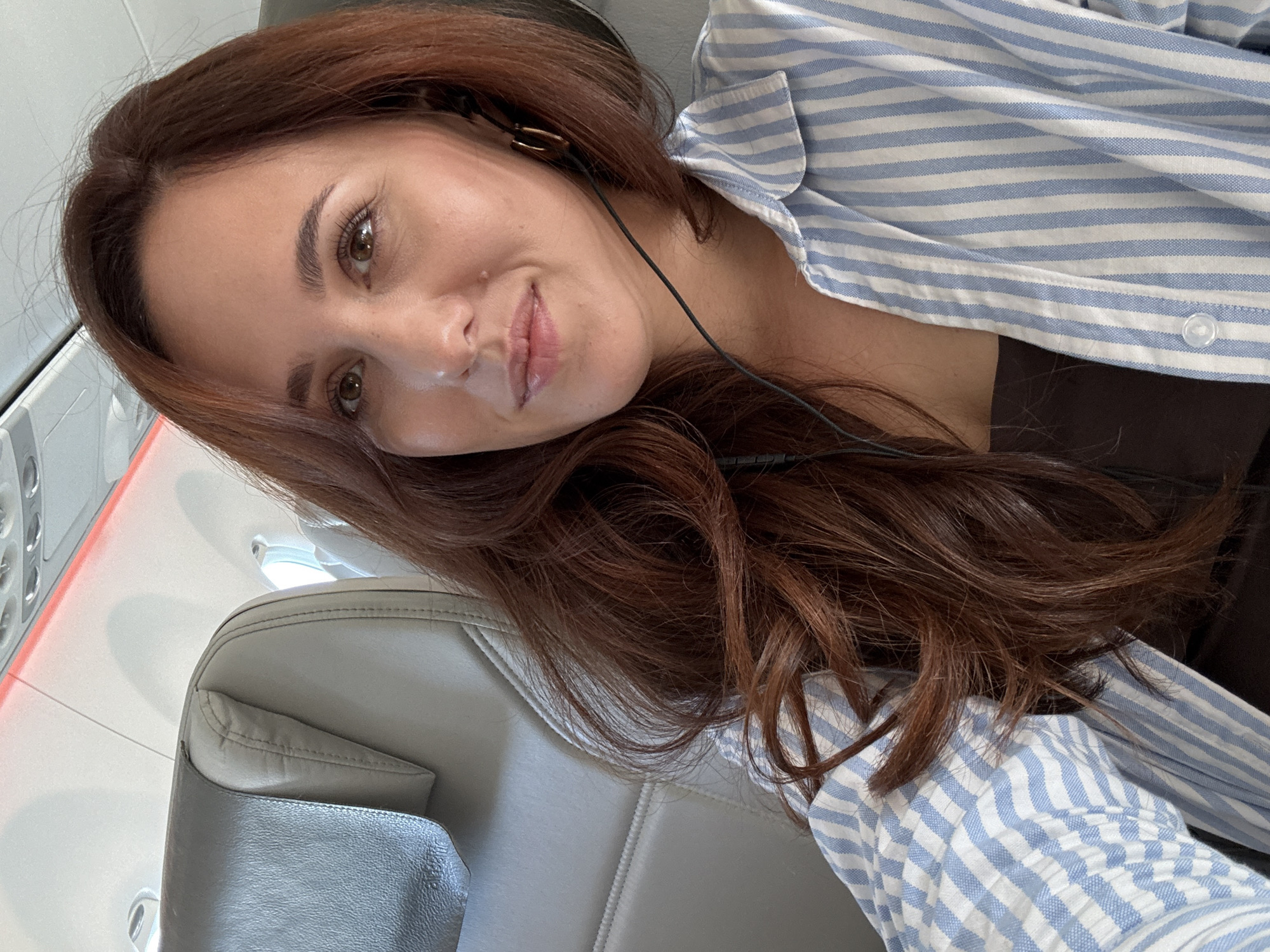 A woman is seated in what appears to be an airplane. She has auburn hair, brown eyes, and is wearing a blue and white striped shirt. She has earbuds in her ears. The airplane seat is gray. The location is not discernible.
