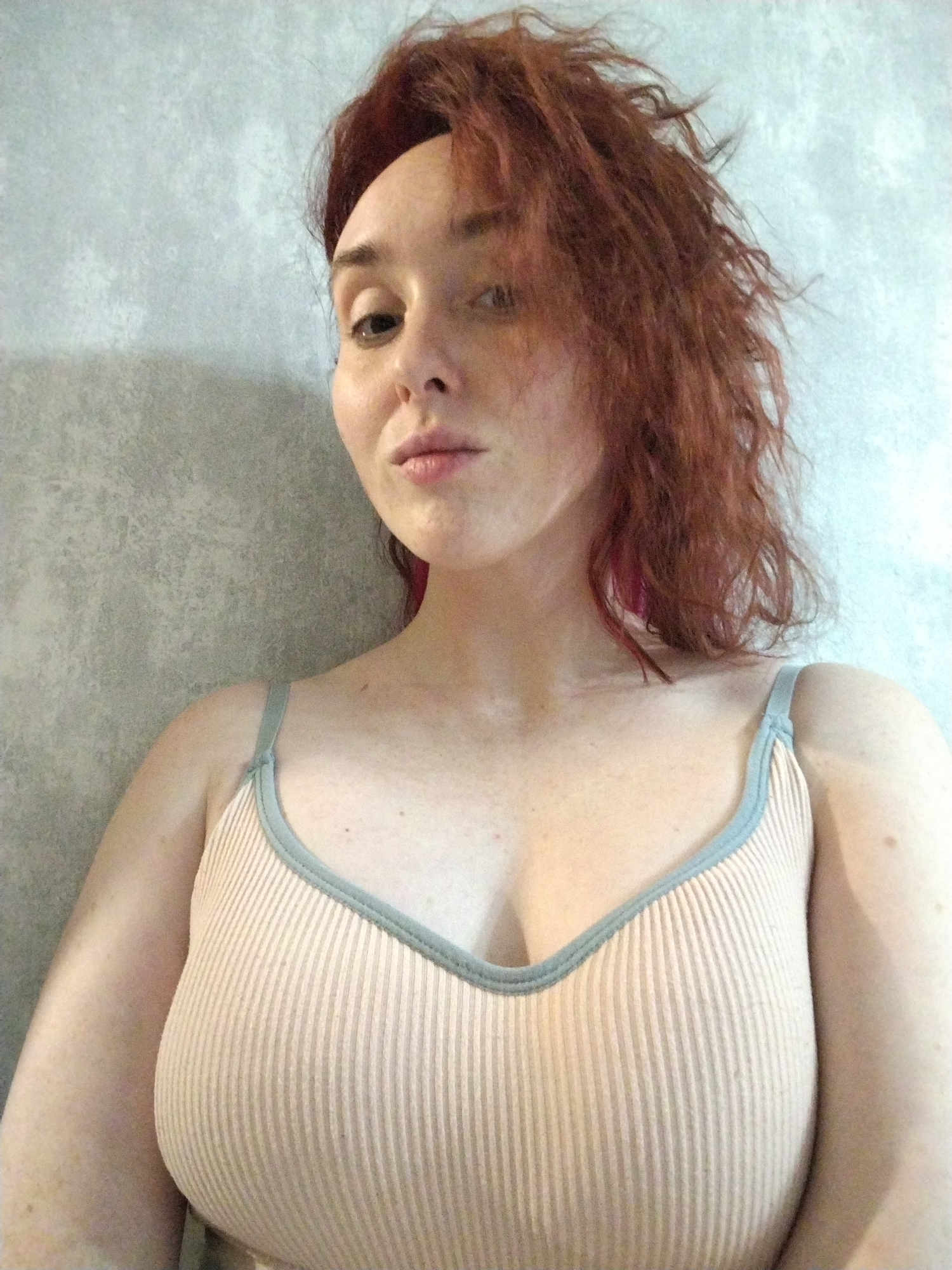A close-up, chest-up portrait features an individual with fair skin, freckles on the décolletage and shoulders, and reddish-brown, dishevelled hair with subtle pink highlights visible on the subject's right side. The subject's face is oriented towards the camera with a neutral expression, direct eye contact, and slightly parted lips. The individual wears a light peach-colored, vertically ribbed tank top with a light blue trim along the neckline and shoulder straps. The background consists of a plain, light grey, textured wall. No specific environmental or contextual details within the image can establish the location as Izhevsk, Russia.