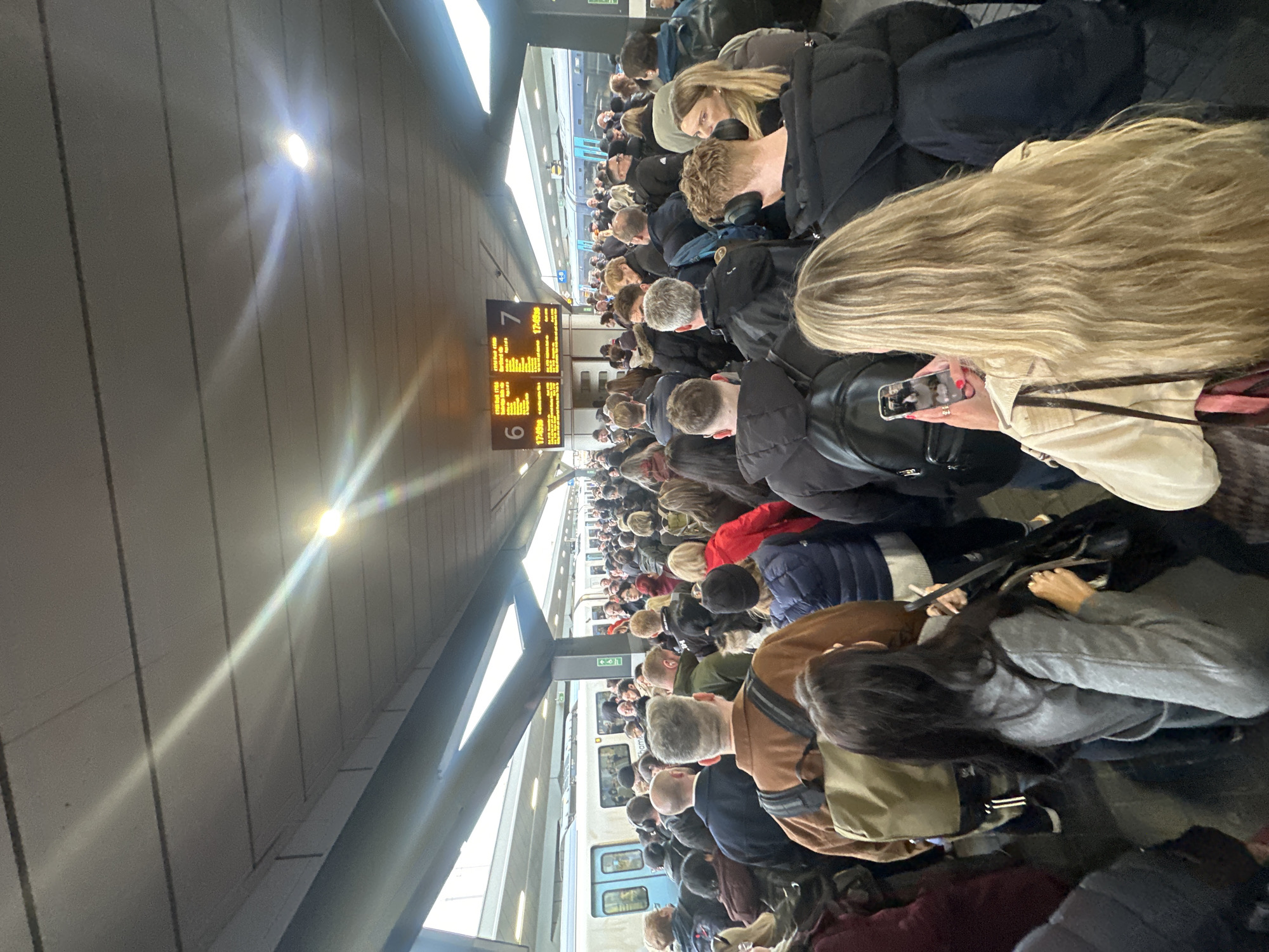 This image depicts a densely crowded train platform at a major station in London, United Kingdom. The train information boards clearly display destinations such as "Farringdon," "City Thameslink," and "St Pancras Intl," indicating a Thameslink line platform.
The scene is an indoor platform under a grey, paneled ceiling with visible linear recessed lighting fixtures, one of which produces a strong lens flare. Two passenger trains, each with a distinctive light blue stripe along their sides, are stationary at adjacent platforms, identified by overhead digital display boards as Platform 6 (left) and Platform 7 (right). The train on Platform 6 has its doors open.
Hundreds of individuals, predominantly adults, are packed tightly together, occupying the entire visible platform space. Most are seen from behind or in partial profile, dressed in various forms of casual to semi-casual winter attire, including coats, jackets, and puffer jackets. They appear to be waiting, with many looking towards the central digital display boards which show train departure times, including "17:49:36". In the immediate foreground, a person with long blonde hair is holding a smartphone horizontally, pointed away from the camera, as if taking a photo or video of the crowd and platform. Other individuals are also holding smartphones, and some wear backpacks or carry bags. One person in the mid-ground wears over-ear headphones.