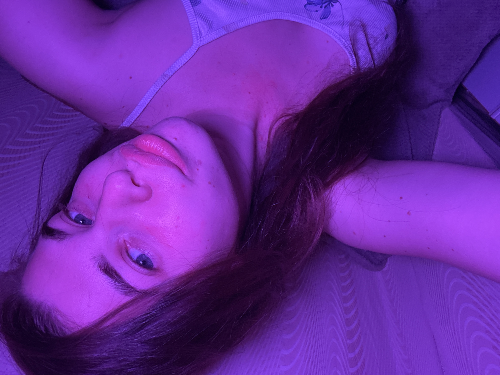 The image shows a close-up, high-angle shot of a young woman lying down, illuminated by purple light. She has long, dark hair that falls across her face and shoulder. Her eyes are open and looking upwards, and her lips are slightly parted. She is wearing a light-colored tank top with a floral pattern. The background appears to be a textured surface, possibly a couch or bed. The overall lighting is atmospheric and casts a strong purple hue over the entire scene. No specific location in Мещёры, Russia can be determined from the image.