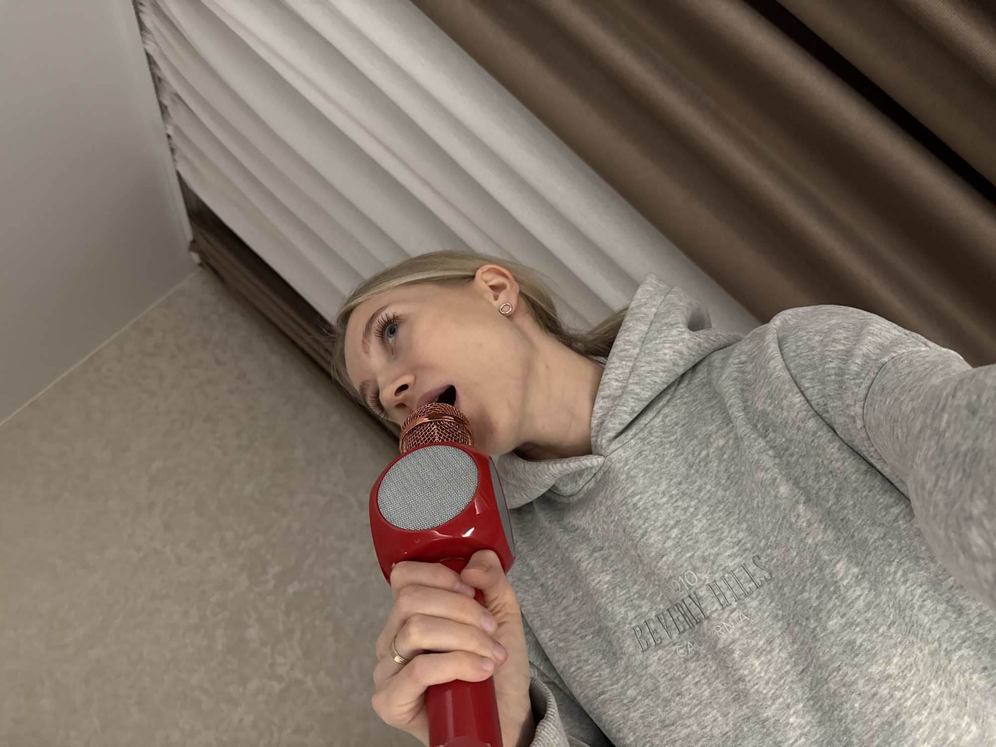 A woman with blonde hair and wearing a grey "Beverly Hills California" hoodie is holding a red microphone with a grey speaker in front of her mouth, as if singing. She wears a simple hoop earring in her left ear and a wedding band on her left ring finger. The background includes cream and brown curtains on the upper right and a beige wall on the left. The location is unknown and cannot be identified as Izhevsk, Russia based on the visual information provided.