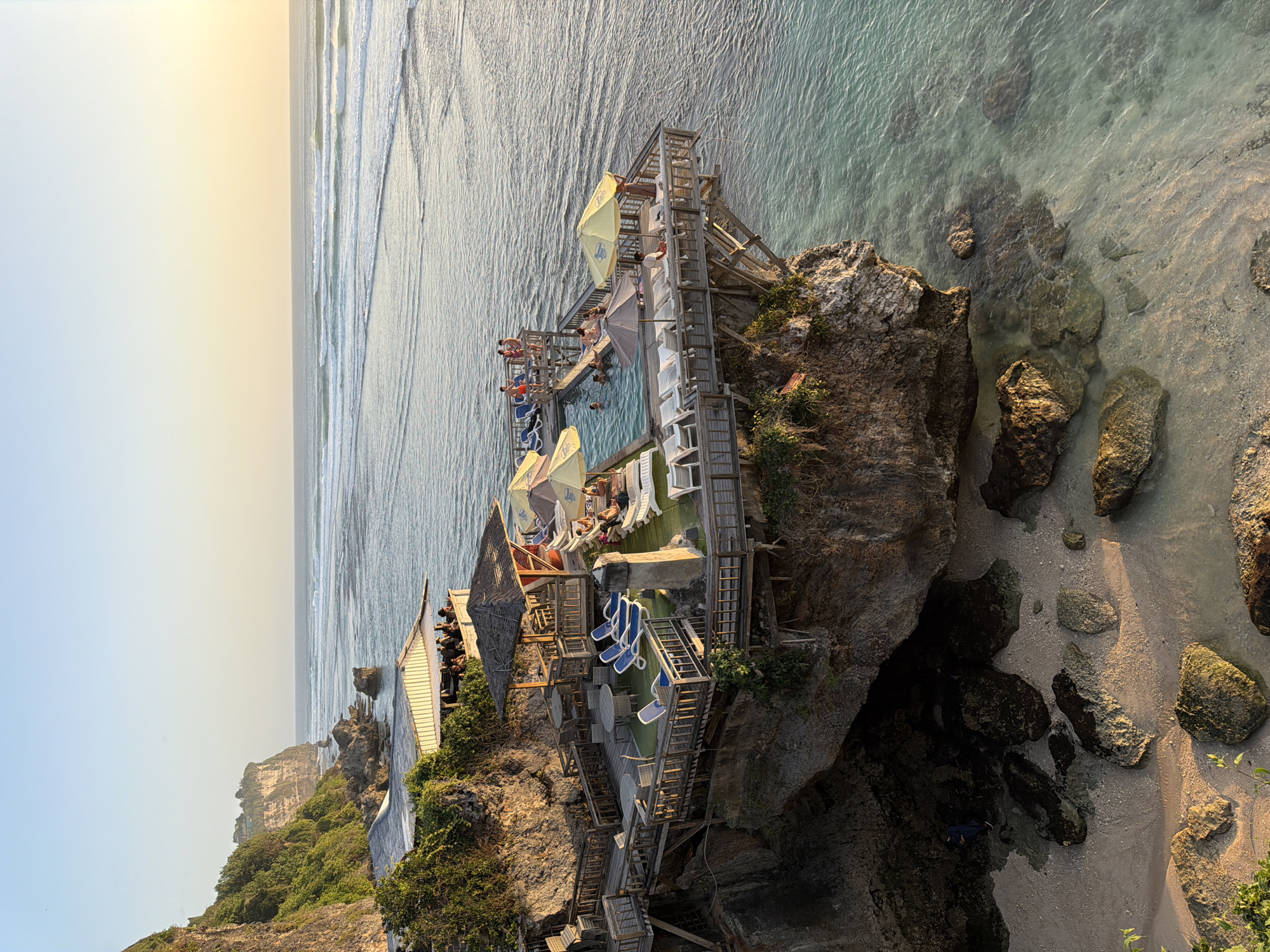 A cliffside restaurant and swimming pool complex overlooks the ocean at sunset in Kuta Selatan, Indonesia. The complex is built on multiple wooden decks extending from the cliff face. The upper deck features a covered restaurant area with patrons seated at tables. Below this, a second deck has several dining tables and lounge chairs. Further down and cantilevered over the water is a swimming pool with several people swimming and lounging. Yellow and grey umbrellas are scattered around the pool area. The ocean in the background has gentle waves. The sun is low in the sky, casting a warm glow. The cliff face is covered in lush green vegetation. Below the complex, a sandy beach with large rocks is visible at the base of the cliff.