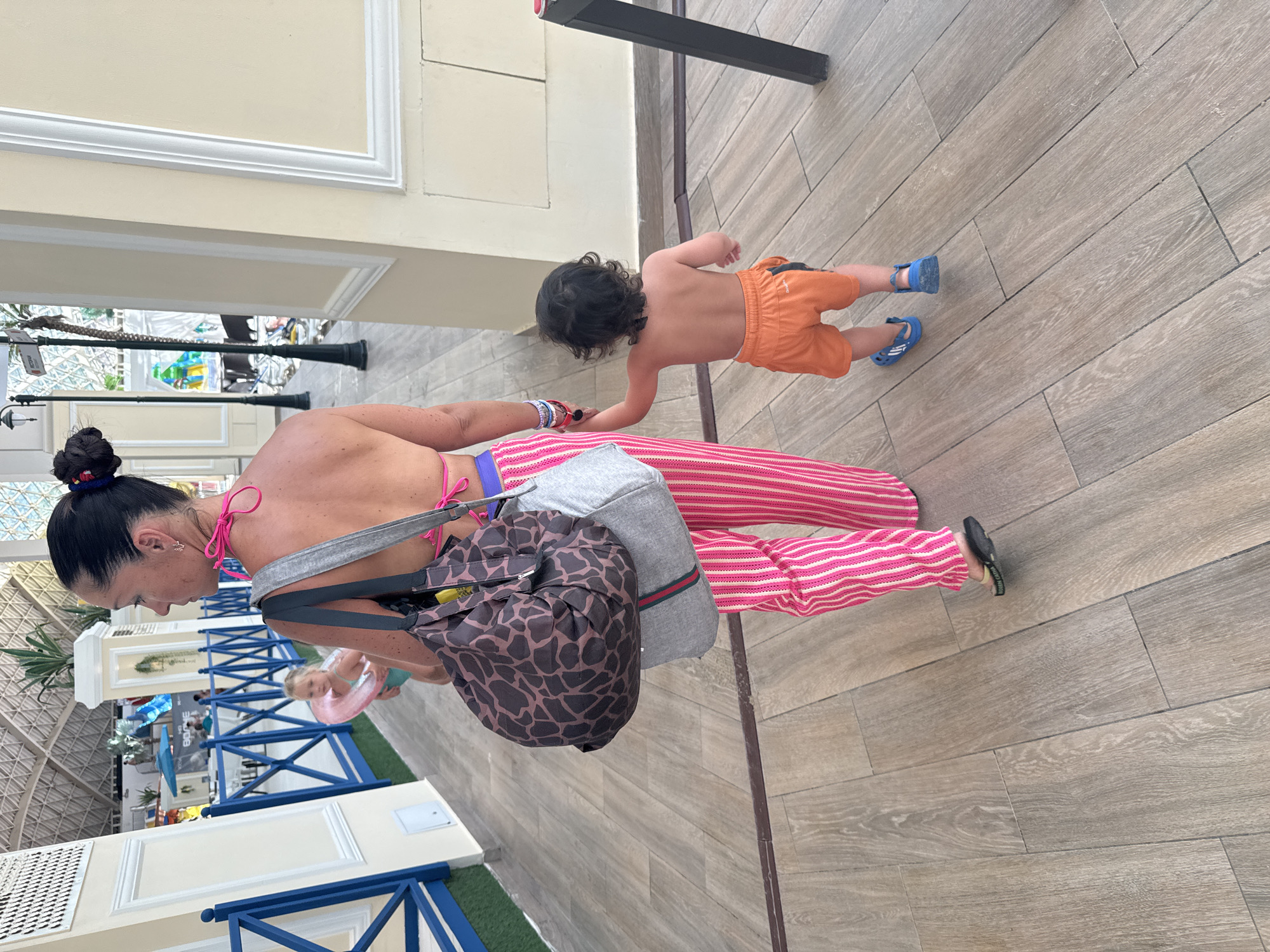 The image shows a woman and a young child walking on a tiled floor. The woman is wearing a pink bikini top and pink and white striped pants. She has a bun in her hair and is carrying a large bag with a giraffe print. The child is wearing orange shorts and blue sandals. The child is holding the woman's hand. In the background, there is a blue railing and some decorative elements. The overall setting appears to be an indoor or covered outdoor area, possibly a resort or a shopping center, given the architecture and tiled flooring. There are no clear indicators of the time of day or weather. The interaction between the woman and child suggests a casual outing, possibly a family vacation.