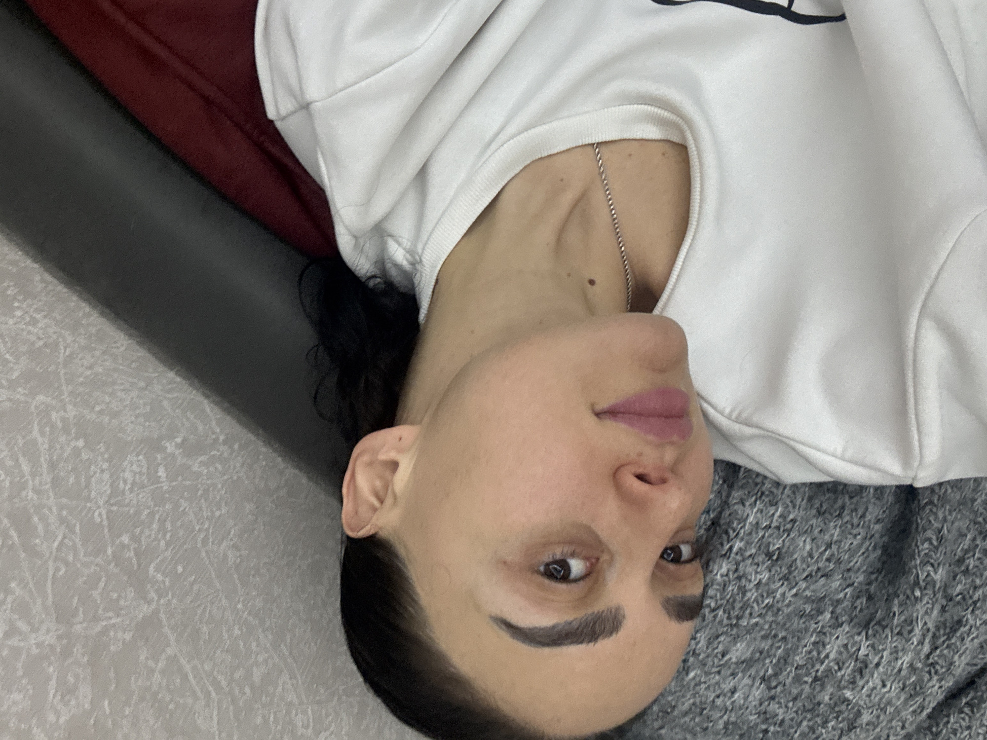 A medium shot of a woman with dark hair lying on a grey blanket, wearing a white sweatshirt and a silver chain. The background consists of a grey wall, a black surface, and a burgundy object. There is no indication of the location being Tbilisi, Georgia.