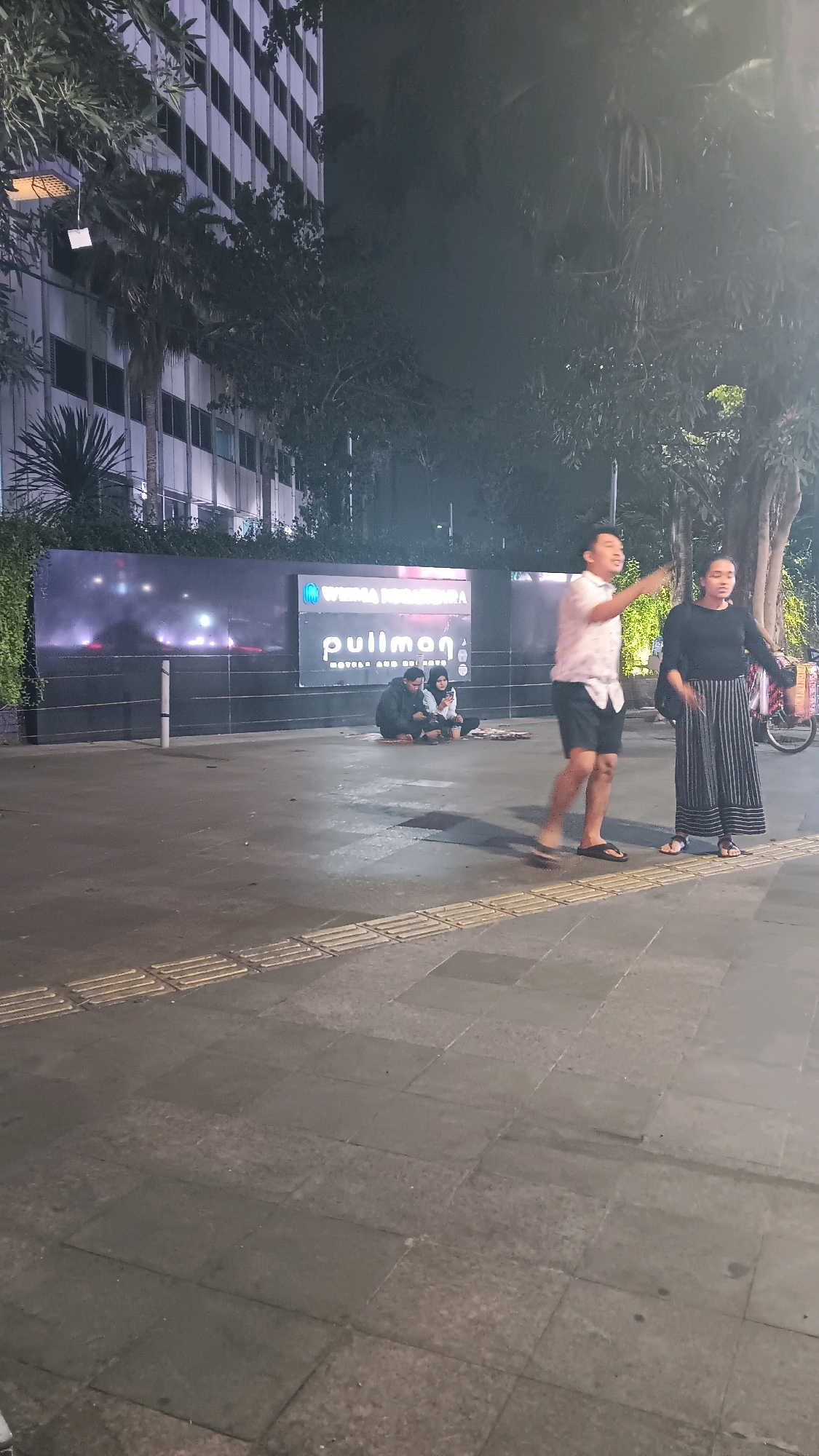 In Central Jakarta, Indonesia, at night, two individuals stand on a paved sidewalk in front of a building with a large "Pullman" sign. The man, wearing a patterned short-sleeved shirt and shorts, gestures with his right hand. The woman beside him, in a black long-sleeved shirt and a striped maxi skirt, stands with her arms outstretched. In the background, two people sit on the ground near a wall. The environment includes trees, building facades, and streetlights.