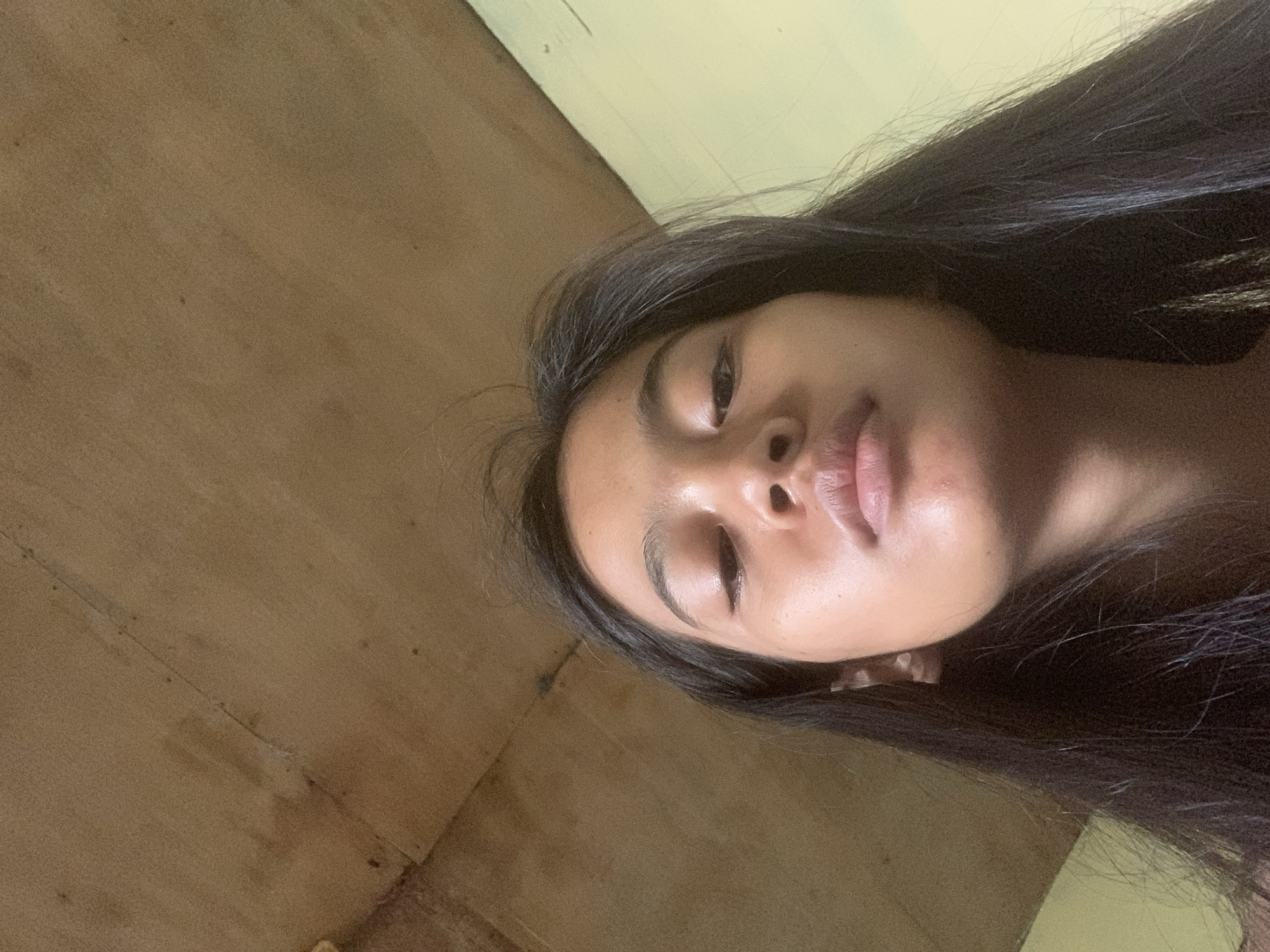 The image is a low-angle, close-up, selfie-style shot of a young woman's face looking downwards. She has long, dark hair that frames her face, and her skin appears smooth and clear. Her eyes are partially closed, and her lips are slightly parted. The background consists of a textured wooden ceiling with visible seams and stains, and a pale yellow wall to the right. The lighting suggests an indoor environment, possibly during the day. The context of Sungai Meriam, Indonesia, is not visually present in the image itself; it is a geographical identifier for the location where the photograph was taken.