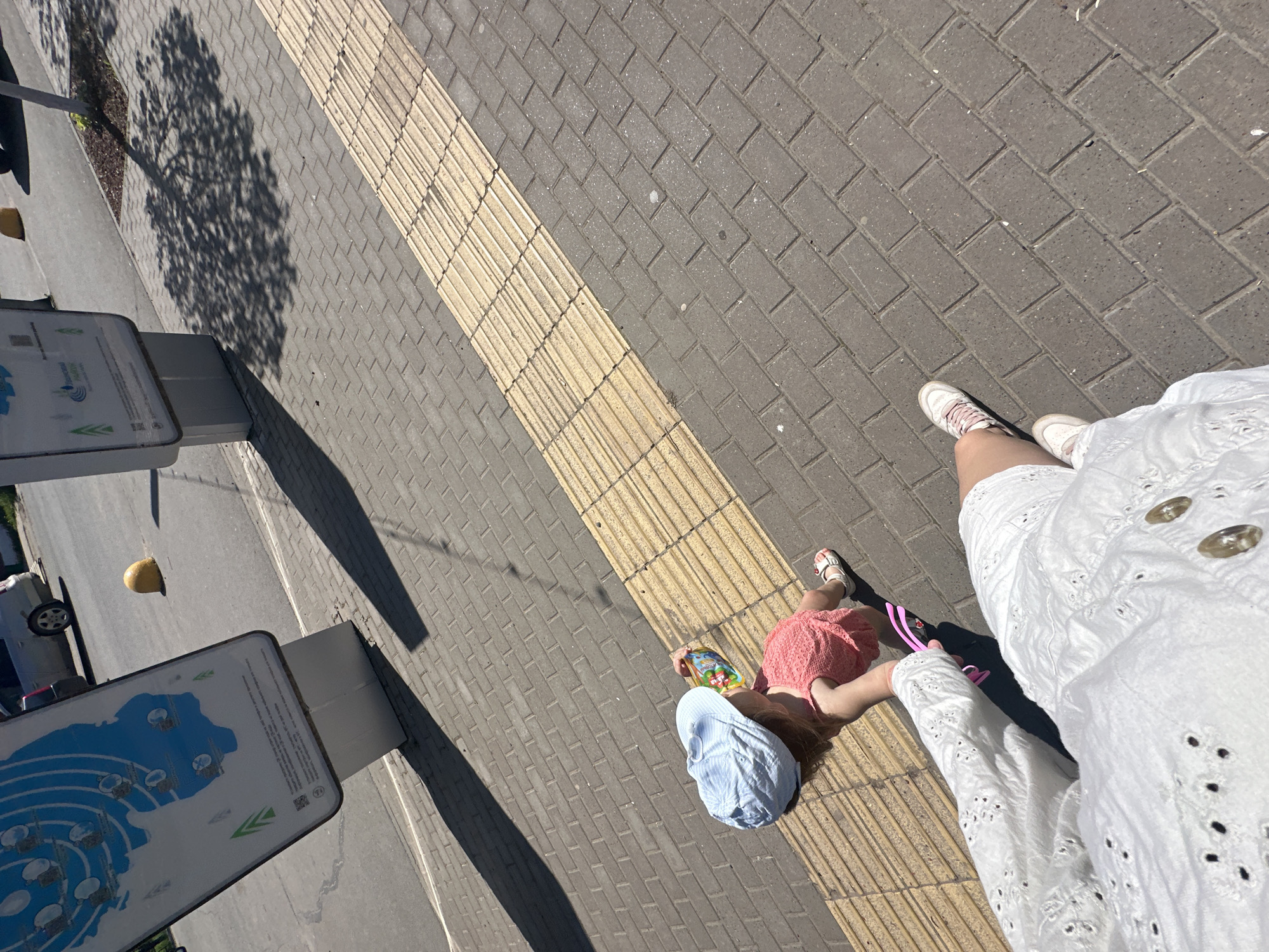 A high-angle shot shows two children lying on a sidewalk. One child wears a light blue hat and holds a book. The second child is next to the first wearing a red dress and sandals. A woman wearing a white dress stands over the children, holding a pink object. A sidewalk with textured yellow stripes lies beneath the children. A tree’s shadow falls on the sidewalk. Two signs are on the left side of the image. The location is in the city of Uva, Russia.