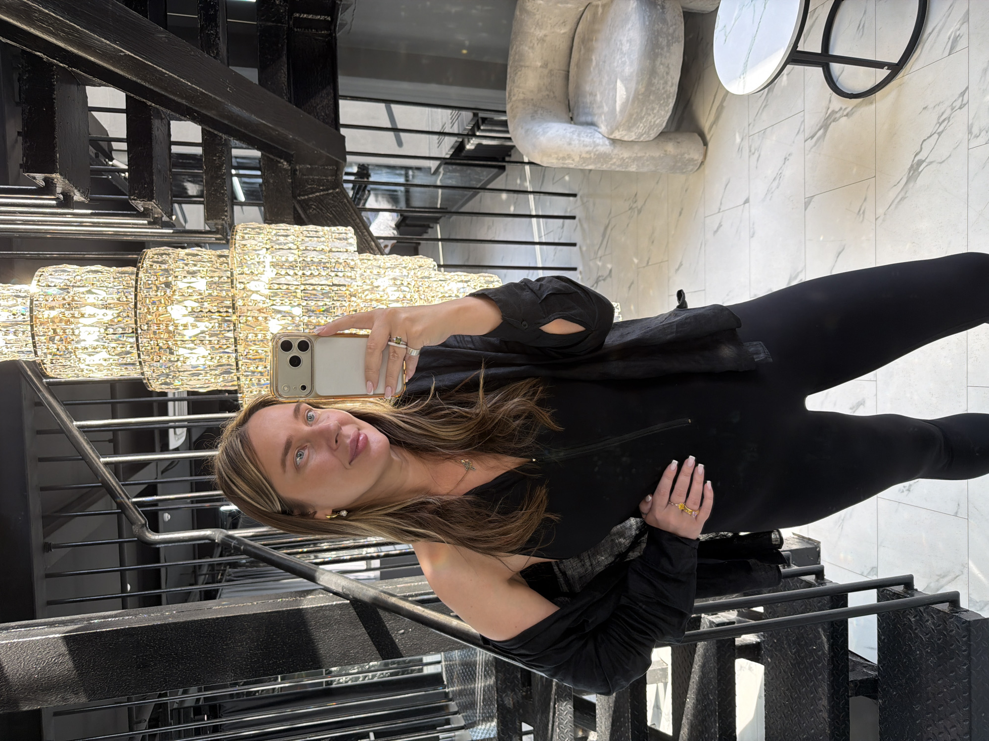 A woman is taking a selfie indoors. She is standing on a black metal staircase, adjacent to a large crystal chandelier. She is wearing a black outfit, and has shoulder-length blonde hair. Her phone is in her right hand, and her left hand is near her waist. In the background, there's a grey chair and a round table on a marble floor. The location is possibly in Moscow, Russia, but there is no definitive evidence in the image to support this.