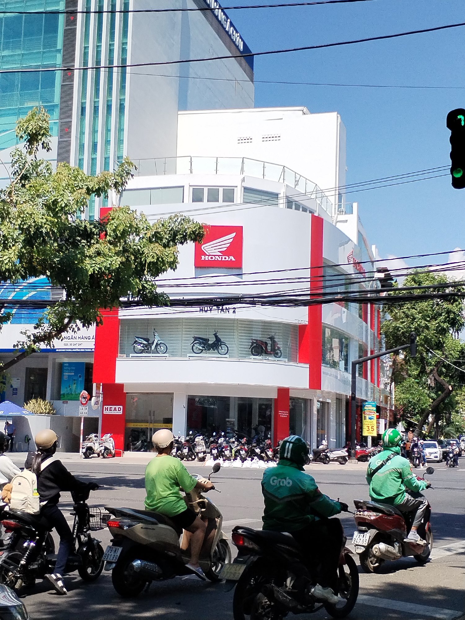 The image shows a street scene in Nha Trang Ward, Vietnam, featuring a Honda dealership. The dealership is a two-story building with a red and white facade, displaying the Honda logo prominently. Several motorbikes are on display in the windows. In front of the dealership, a group of people on motorbikes are waiting at a crosswalk, with a green traffic light visible above them. Two riders are wearing green Grab delivery uniforms. A bank is seen in the adjacent building. Power lines are visible overhead, and a mix of modern buildings form the urban backdrop. Street signs and traffic markings are present. Vehicle license plates are visible.