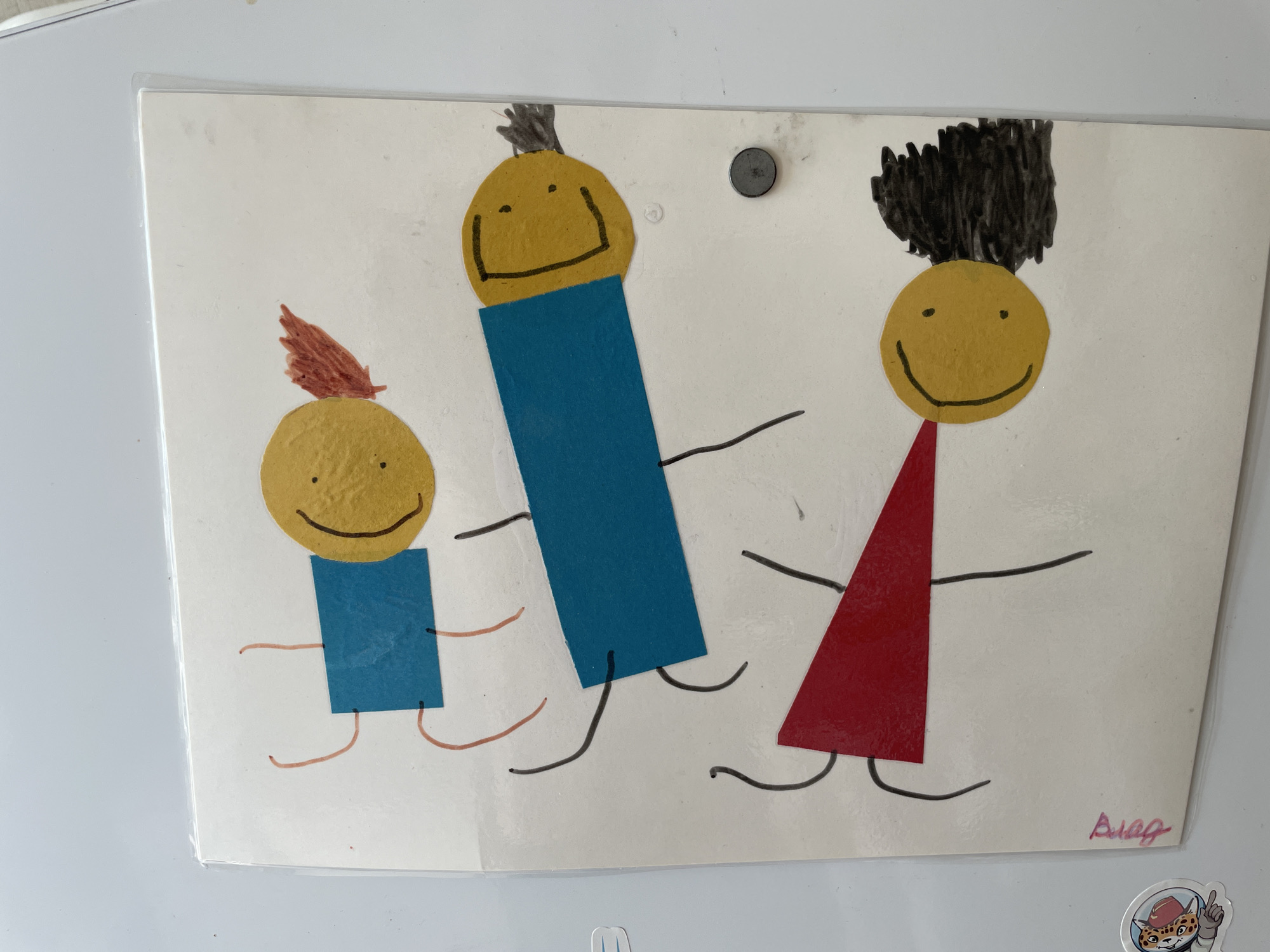 This is a close-up, eye-level, indoor photograph of a child's drawing attached to a white surface, possibly a refrigerator, with a magnetic clip. The drawing depicts three stick figures with yellow circular heads and simple black facial features (eyes and mouths). Each figure has a colored body; the smallest figure on the left has a blue rectangular body and a red feather-like tuft of hair. The middle figure is taller with a long blue rectangular body. The tallest figure on the right has a red triangular body and dark, spiky hair. All figures have thin black lines for arms and legs. A small round grey magnet is visible near the top center of the drawing. In the bottom right corner of the drawing, there is a pink scribbled signature. A small sticker of a cartoon leopard in a spacesuit is visible below the drawing. The drawing appears to be located in a domestic setting, potentially in the city of Laishevo, Russia.