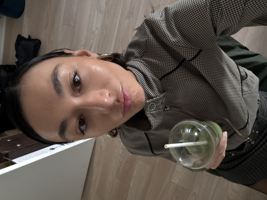 A female individual is depicted in a close-up, selfie-style photograph within an indoor setting. The subject has dark, slicked-back hair, a medium complexion, dark eyes, and glossy lips in a slight pout. She wears large gold-toned hoop earrings and a brown and black small-checkered jacket with a high collar, a zipper closure, and black piping. A dark garment is visible below the jacket. In her right hand, she holds a clear plastic cup containing a green liquid with a white straw. The background consists of light-colored wooden floorboards. A partial white vertical surface on the left displays inverted, partially obscured text resembling "Laasen." Indistinct dark objects are visible in the upper right background. No discernible features within the image indicate the specific city of Paris, France.