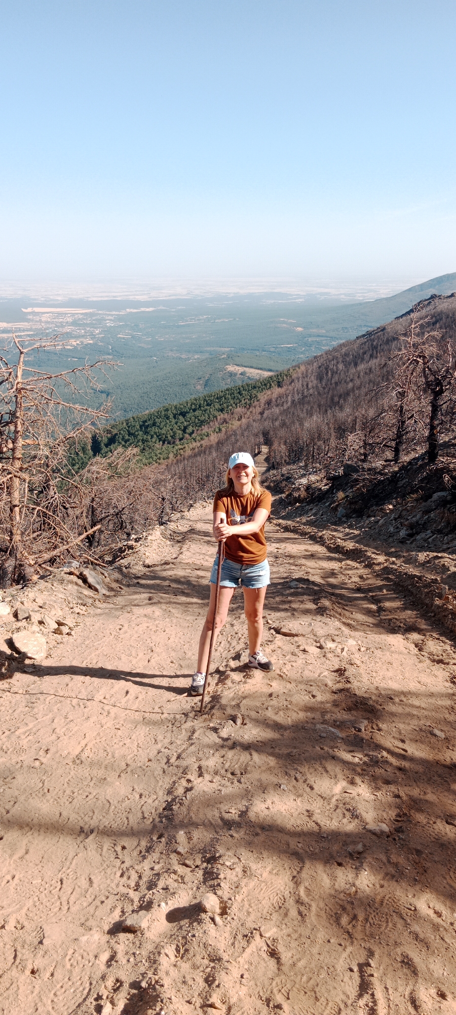 A woman stands on a dirt path on a mountainside in Cerezo de Arriba, Spain, holding a walking stick. She is wearing a rust-colored t-shirt, denim shorts, a light blue baseball cap, and athletic shoes. The path is sandy and worn, with tire tracks visible. To her left, there are dead, bare trees, likely remnants of a wildfire, with dry branches. To her right, the slope of the mountain is covered in dark, scorched trees, indicating further fire damage. Beyond the damaged trees, a dense green forest stretches down the mountainside, eventually giving way to a hazy, distant landscape of fields and a flat horizon under a clear blue sky. The woman is looking at the camera and smiling.