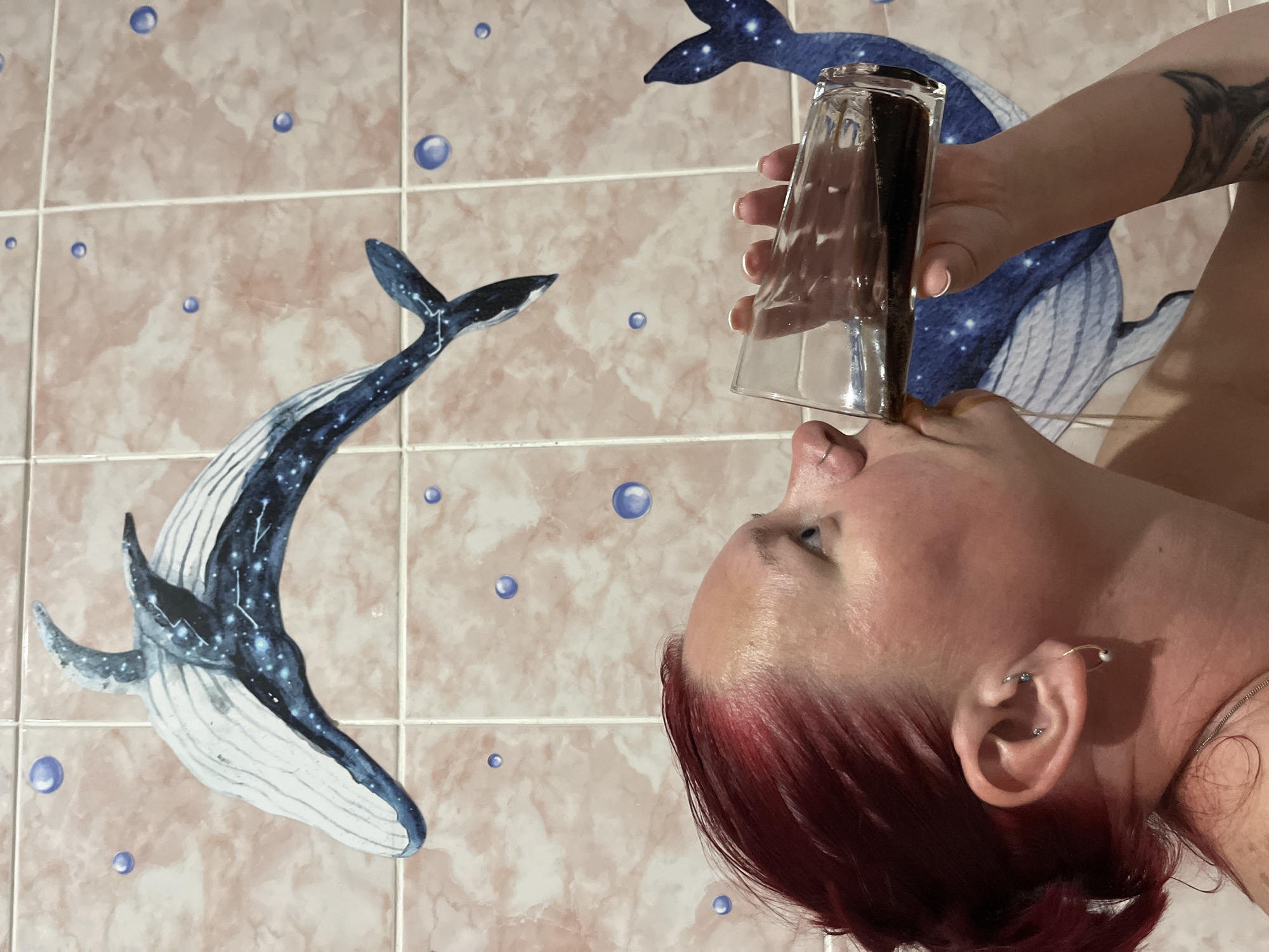A woman with red hair is tilting a glass of dark liquid towards her nose. The image is oriented so the background is oriented vertically. The background features tiled walls with a whale and bubble motif. The location is Chaikovsky, Russia.