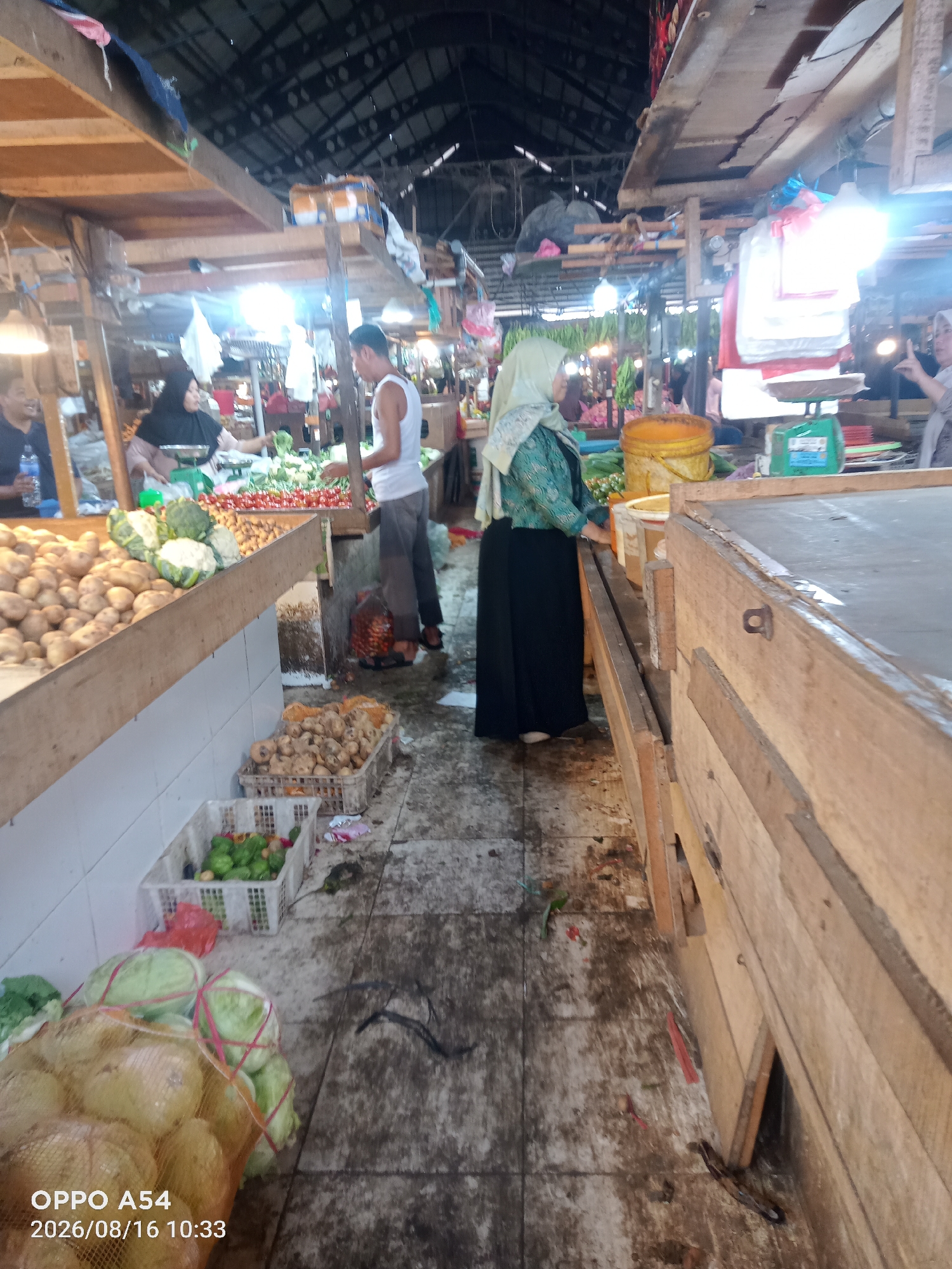 The image depicts an indoor market scene, likely in Jambi, Indonesia, characterized by wooden stalls and a corrugated metal roof. In the foreground, a large mesh bag filled with light yellow produce, possibly onions or gourds, rests on a tiled floor that appears wet and somewhat dirty, with scattered debris. To the left of this bag, two white plastic baskets contain green vegetables, some appearing to be limes or similar fruits, and other leafy greens. Further left, a wooden counter is laden with potatoes, broccoli, and cauliflower.
In the middle ground, a man in a white tank top and grey pants stands near the counter, facing towards the left. To his left, a woman wearing a black hijab and a patterned dress is visible behind a scale, seemingly engaged in a transaction. Another woman, wearing a light green hijab and a patterned blue and green top over a long black skirt, stands to the right of the counter, facing the produce. Behind her, a large yellow bucket and other market supplies are visible. The background shows more stalls and produce, including bunches of bananas hanging. The overall impression is a busy, functional marketplace. The timestamp "OPPO A54 2026/08/16 10:33" is present in the bottom left corner.