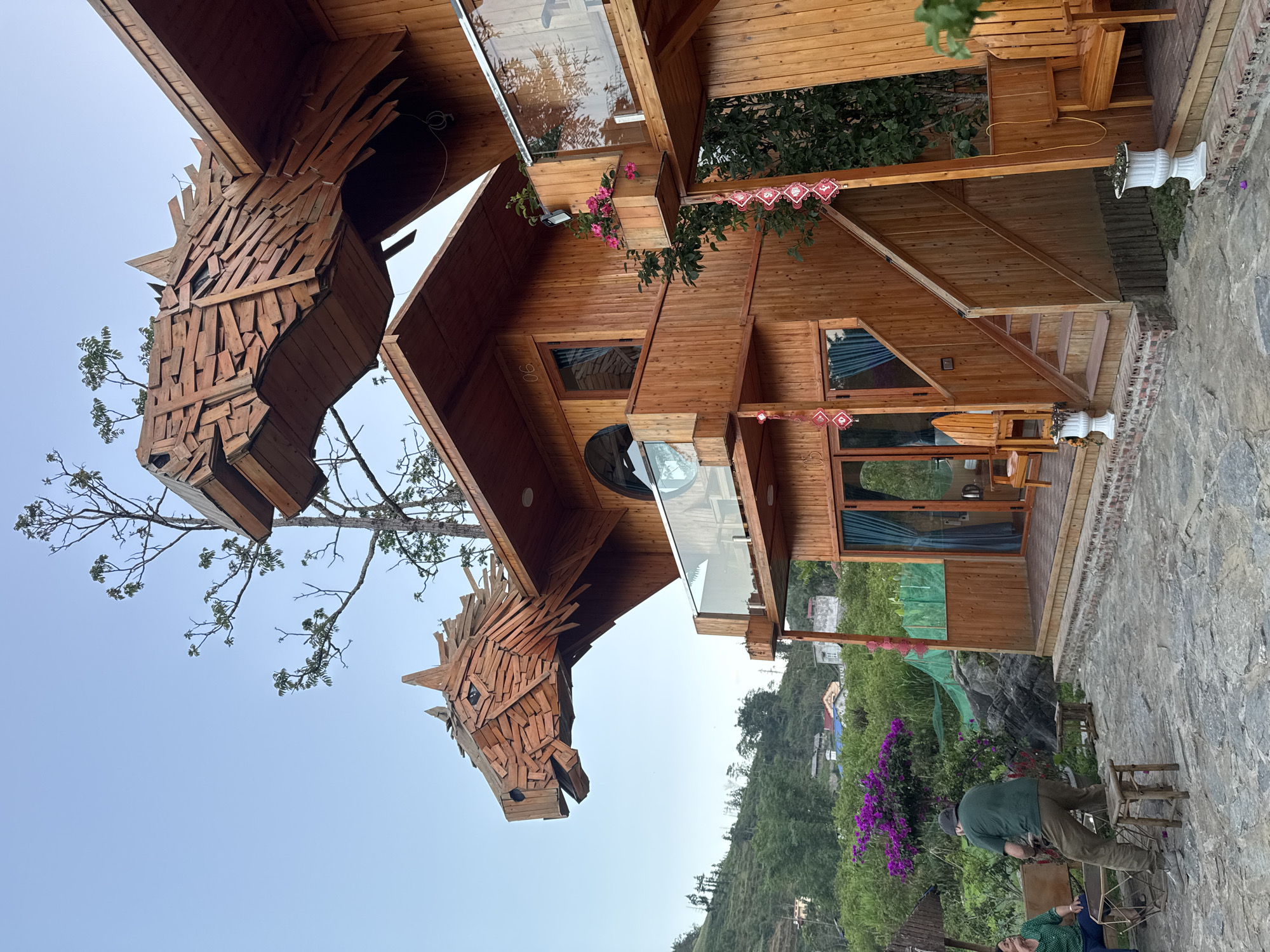 The image depicts a wooden house in Bắc Hà, Vietnam, adorned with two large, carved wooden horse heads protruding from the roof. The house itself is constructed primarily of wood, featuring windows, balconies, and traditional Vietnamese decorations. In the foreground, two people are sitting on chairs, while the background features lush green hills and other buildings. The sky is a clear blue.