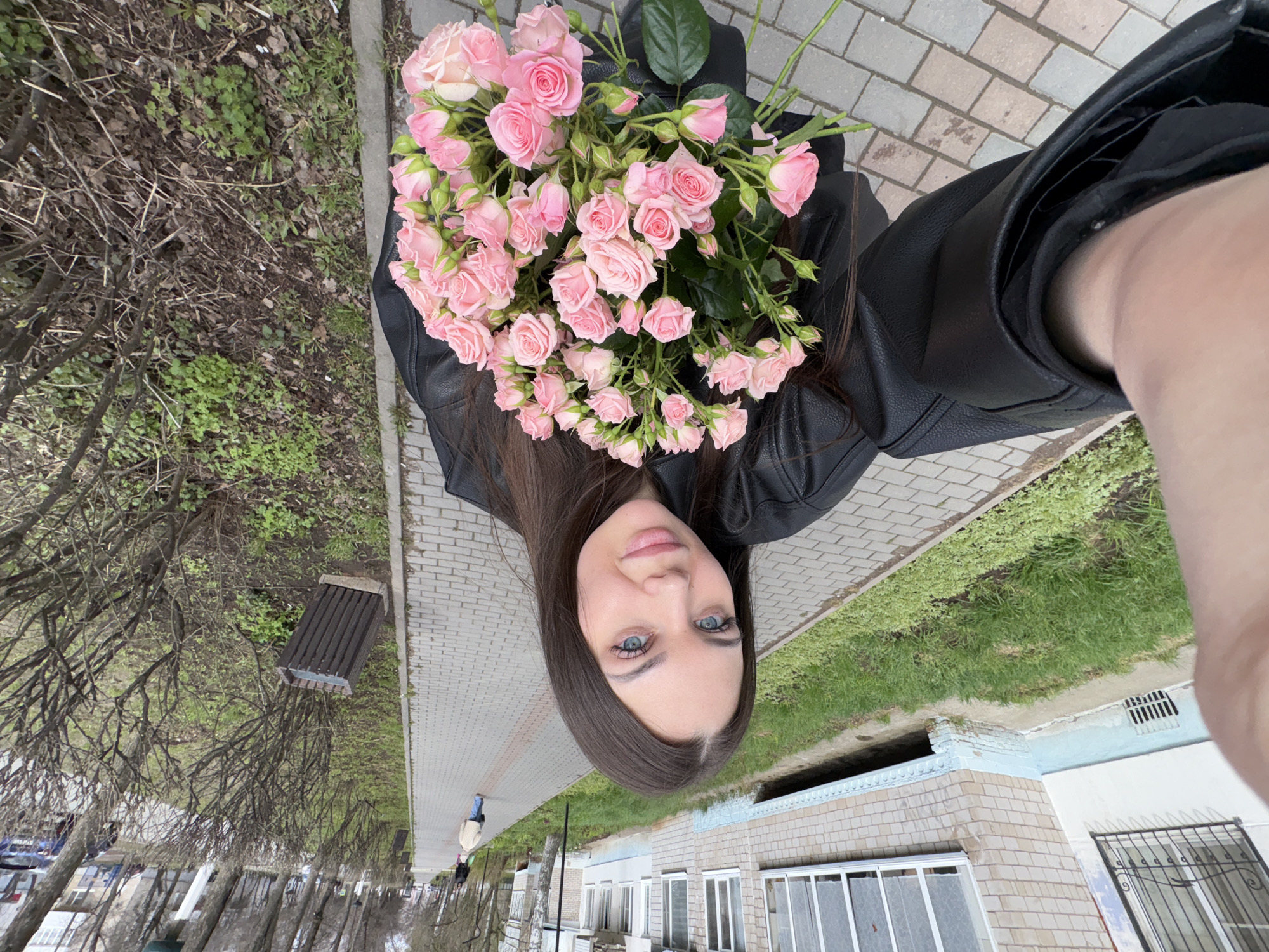 A young woman with long dark brown hair and striking blue eyes poses for a selfie, holding a large bouquet of light pink spray roses. She is wearing a black leather jacket and looks directly at the camera with a neutral to slightly pleasant expression. The setting is an outdoor urban or suburban environment, consistent with Chaikovsky, Russia, during what appears to be late winter or early spring.
She stands on a paved sidewalk made of interlocking grey and reddish-brown bricks. To her left, beyond a strip of green grass and some low shrubs, are numerous bare trees, their branches devoid of leaves. Further down this path, a wooden slatted bench is visible. To her right, several buildings line the street; one distinctive building features light-colored brickwork with a light blue painted base or trim, and some of its windows are fitted with black metal security grates. The time of day is daytime, possibly overcast, given the soft, even lighting and lack of strong shadows. No visible text is present in the image.