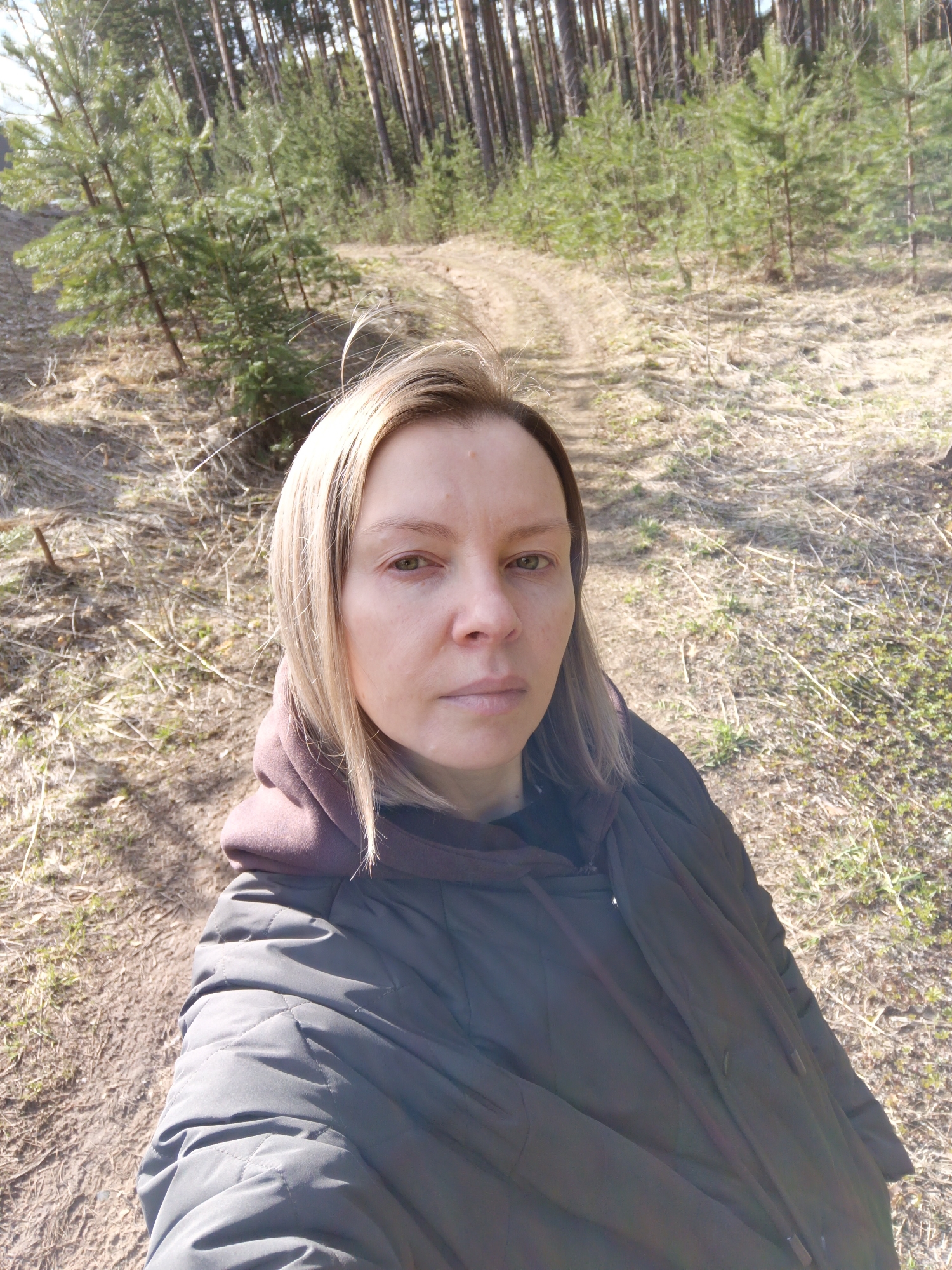 A woman takes a close-up selfie outdoors, facing the camera with a neutral expression. She has fair skin, light brown or blonde shoulder-length hair, and appears to be in her 30s or 40s. She is wearing a dark, possibly black or very dark brown, puffer jacket with a hood, over what seems to be a reddish-brown hooded sweatshirt.
The setting is a sunlit forest, characteristic of regions like Chaikovsky, Russia, as specified. Behind her, a dirt path or track winds gently into the background, flanked by various types of coniferous trees. Young, vibrant green spruce or pine saplings are scattered in the mid-ground, while taller, mature pine trees with straight, light-colored trunks form a denser forest further back. The ground is covered with dry, light-colored grass and fallen pine needles, with sparse patches of new green growth emerging, indicating an early spring season.
The time of day is sunny, with bright, clear light casting visible shadows, highlighting the textured ground and the woman's face. The overall atmosphere is natural and tranquil, capturing a moment during what appears to be a walk in the woods. There is no visible text in the image.