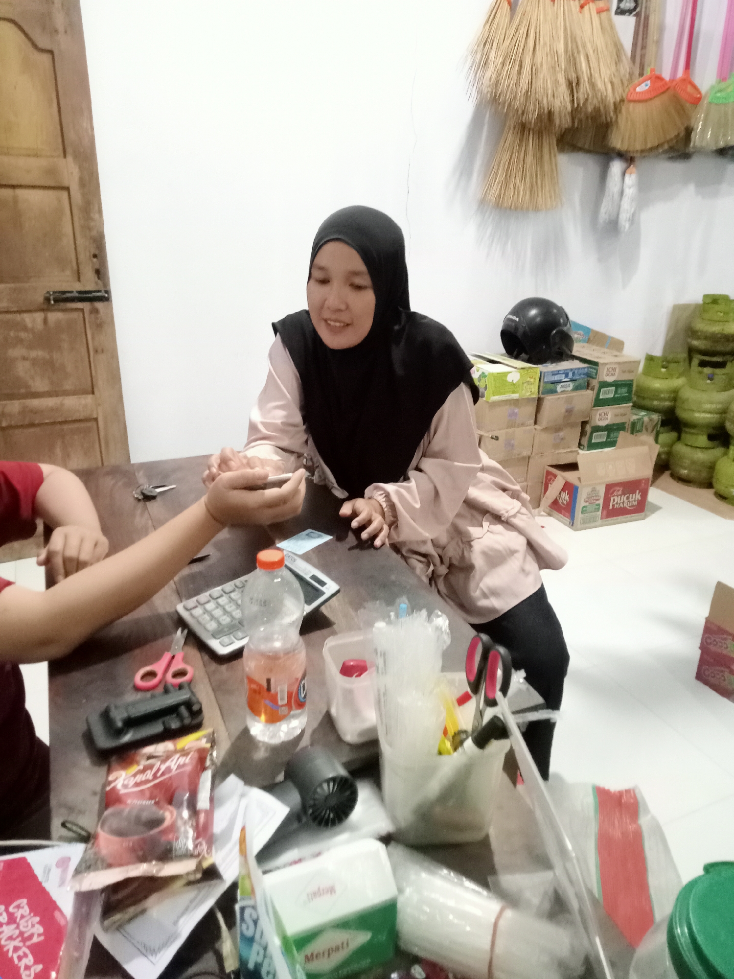The image depicts an indoor scene, likely a shop or a storage area, in Simo, Indonesia. A woman wearing a black hijab and a pale pink long-sleeved top is seated at a wooden table, looking downwards with a slight smile. She is interacting with an unseen person whose arm is visible on the left side of the frame, holding what appears to be a small item. The table is cluttered with various objects, including a calculator, scissors, a bottle of pink liquid, a packet of "Kapal Api" coffee, and other packaging. In the background, stacks of cardboard boxes are visible, some with labels such as "AQUA" and "Pucuk Harum". Several green gas cylinders are also present. The wall behind the woman is adorned with several brooms of different sizes and colors. A dark-colored motorcycle helmet rests on top of the boxes. The overall impression is one of a busy, functional space where transactions or administrative tasks might be taking place.