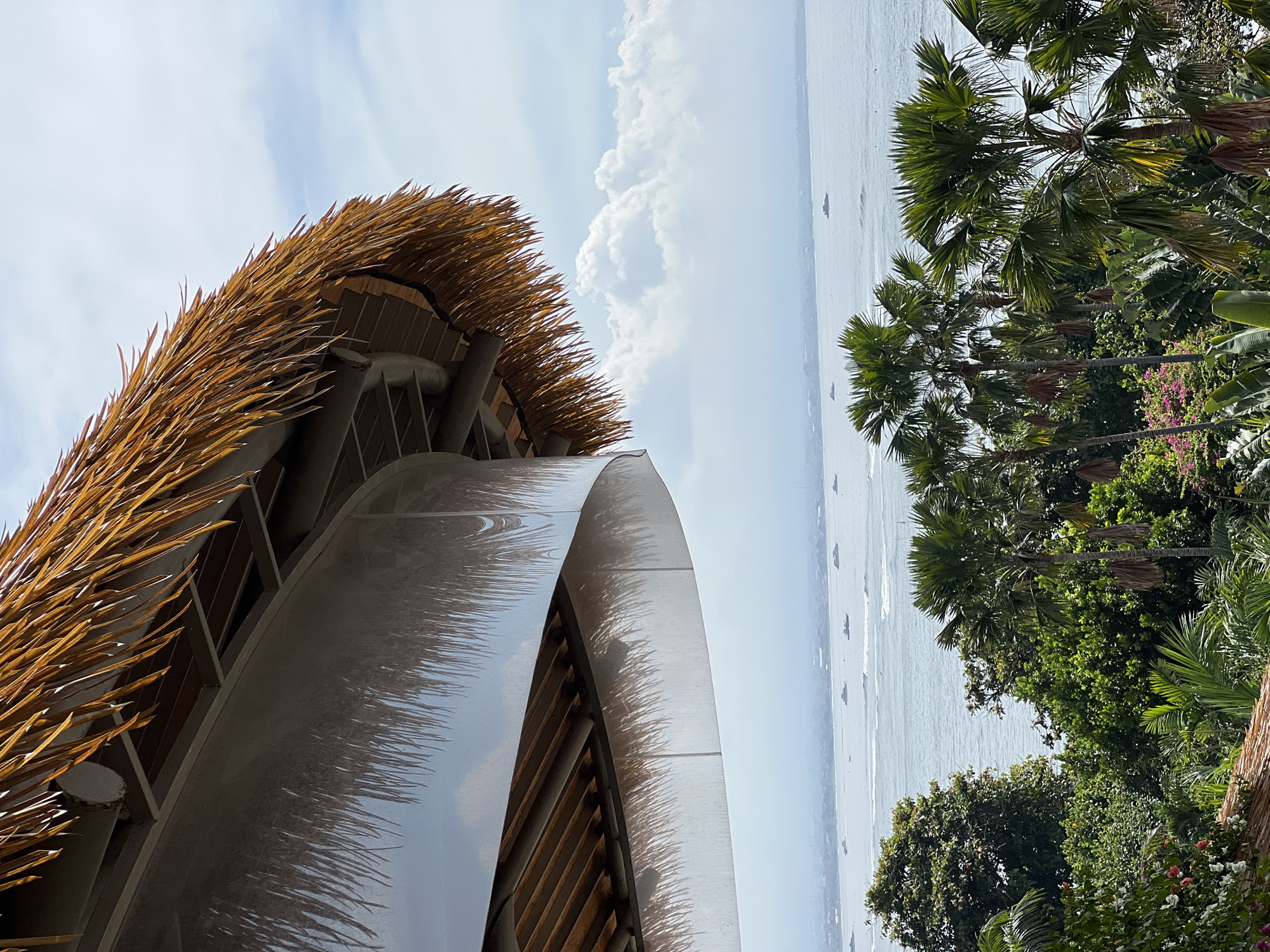 This image, captured in Denpasar, Indonesia, presents a vibrant tropical coastal landscape from a low-angle perspective. The foreground is dominated by modern architecture, featuring a building with a distinctive, intricately layered thatched roof made of dried palm fronds or straw, which exhibits a warm, golden-brown hue. Adjacent to this, a large, smooth, and curved architectural surface, possibly made of metal or polished concrete, reflects elements of the thatched roof and the bright sky, creating dynamic patterns.
To the right, lush tropical vegetation thrives, including several tall palm trees with expansive green fronds and dense clusters of other green trees and bushes, many adorned with small pink and white flowers. Beyond the verdant foliage and architectural structures, a serene expanse of the ocean or sea is visible under a bright, mostly clear sky. Scattered white cumulus clouds drift across the upper part of the sky. Several small boats or ships are discernible in the distance on the water, indicating marine activity.
The scene is bathed in bright daylight, suggesting a clear day during midday or early afternoon. The combined elements create an atmosphere typical of a tropical resort or a high-end coastal establishment. There is no visible text in the image.