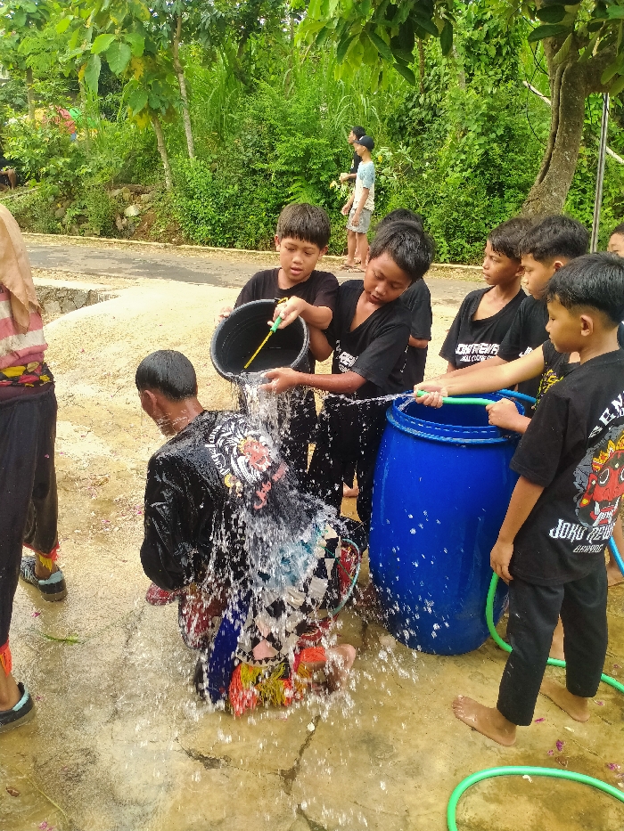 In Gebyog, Indonesia, an outdoor scene captures a group of individuals engaging in a water activity. A central figure, dressed in a black long-sleeved shirt with a prominent, colorful graphic on the back and patterned lower garments, kneels on a wet, light-colored paved surface. Water cascades over this individual from a black plastic bucket, held by a child to their left. Another child to the right operates a green hose, directing water towards the central figure and a large blue plastic barrel. Several other children, all wearing black t-shirts and dark pants, stand nearby, observing or participating in the water distribution from the barrel. Some children are barefoot, while others wear sandals. The immediate ground is wet and features scattered organic debris. The background consists of dense green foliage, including trees and bushes, with a partial view of a road or path. The scene is illuminated by natural daylight.