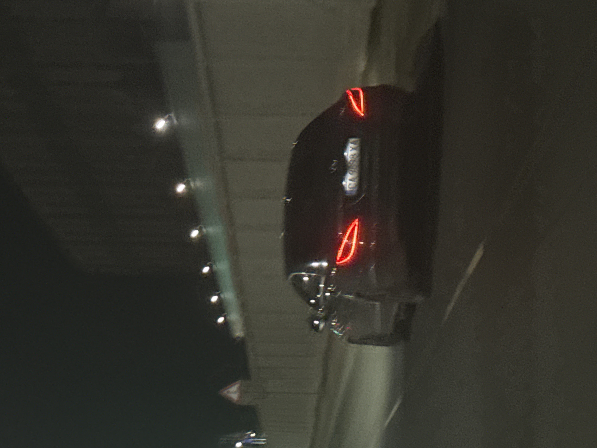 At night, under a bridge or overpass in Plovdiv, Bulgaria, the rear of a dark-colored car is visible. The car's taillights are illuminated, with a distinctive teardrop shape. The license plate is partially visible, with "CA 058 YA" discernible. Above the car, a series of lights are lined up, possibly illuminating the bridge or overpass. The environment is dark and seems industrial or urban. A second car is visible in the background, further away.