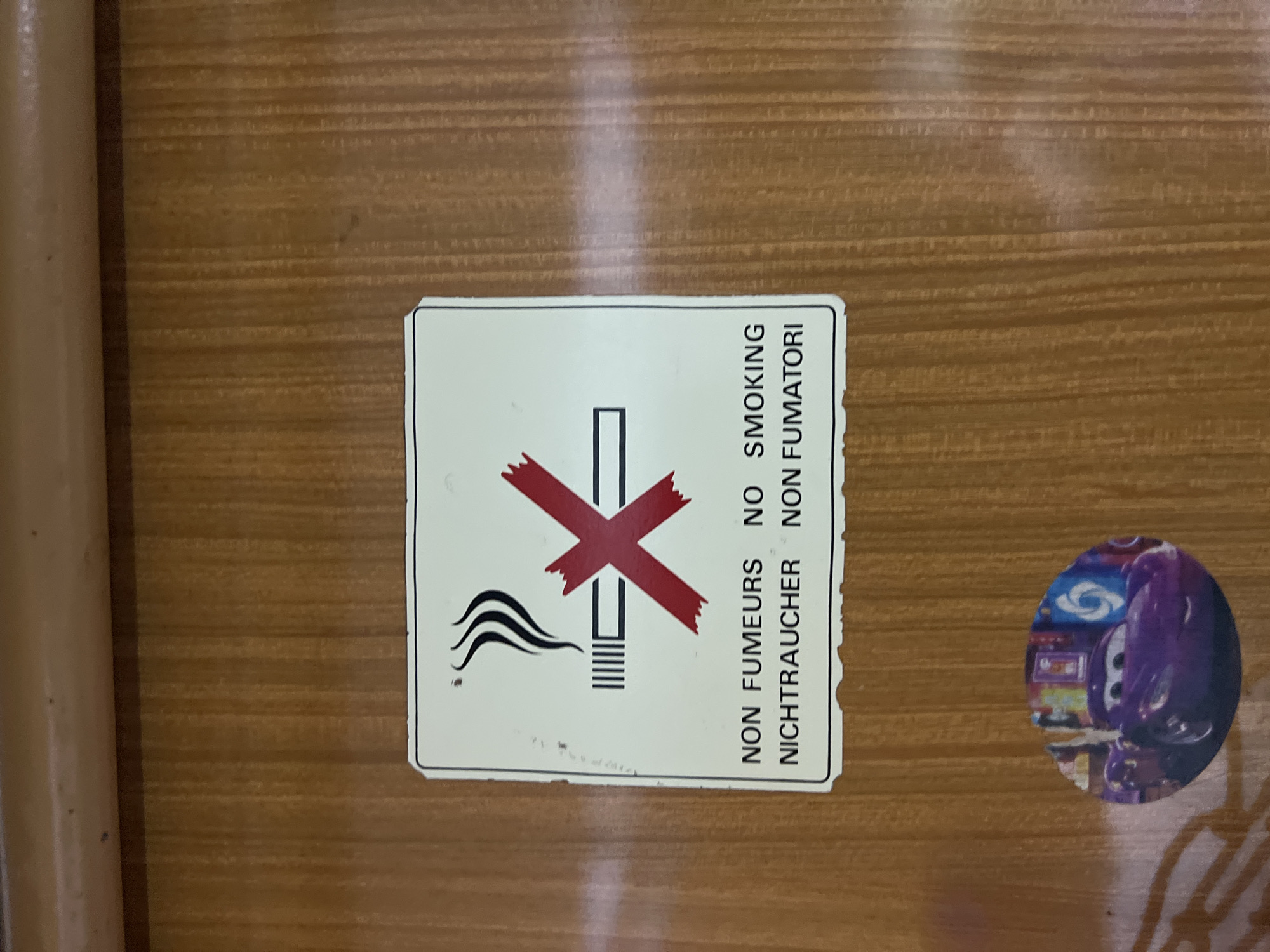 The image shows a "No Smoking" sign with corresponding translations in French, German, and Italian. The sign is rectangular, white with a black border. It has a cigarette symbol with a red "X" over it, along with wavy lines representing smoke. Below the symbol are the words "NON FUMEURS" "NICHTRAUCHER" "NON FUMATORI". To the right of the sign is a small round sticker featuring a purple car from the Pixar movie "Cars". The background is a light brown surface with a wood grain pattern. There is insufficient information to determine if the image was taken in the city of Gornje Mrke, Montenegro. The image is a close-up shot of these items, obscuring the wider environment.