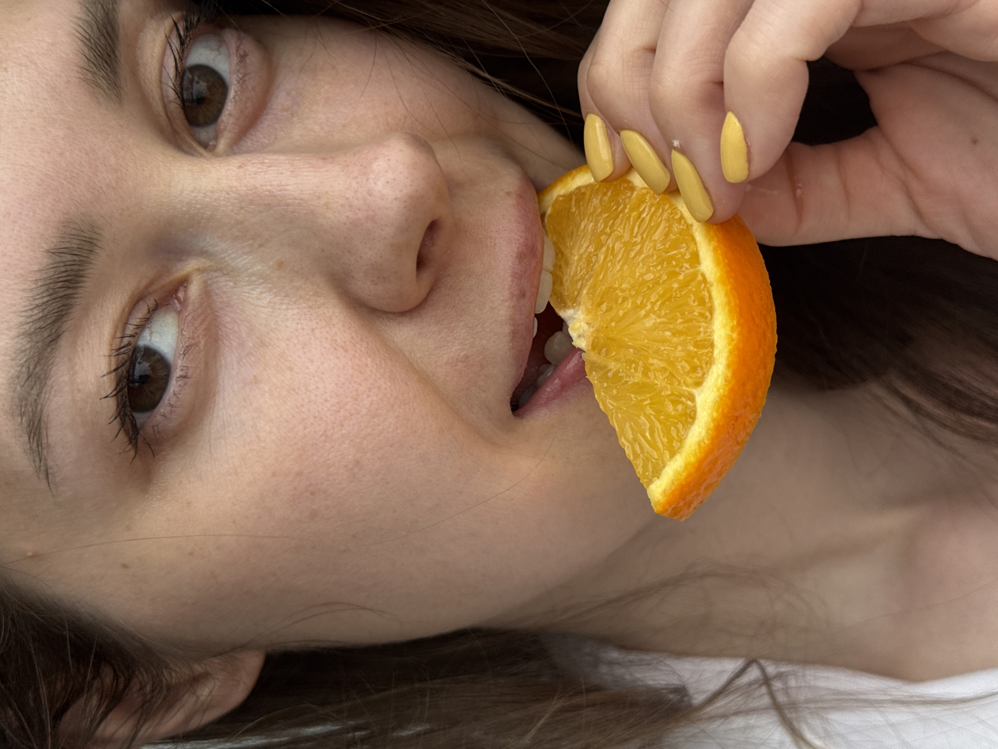 A woman with light skin, brown hair, and brown eyes is holding a slice of orange to her mouth with her right hand. She is lying on her back, with her head turned to the left. Her fingernails are painted yellow. The woman's skin has a few small freckles. A white shirt is visible in the background. There is no evidence to suggest the location is Chaikovsky, Russia.