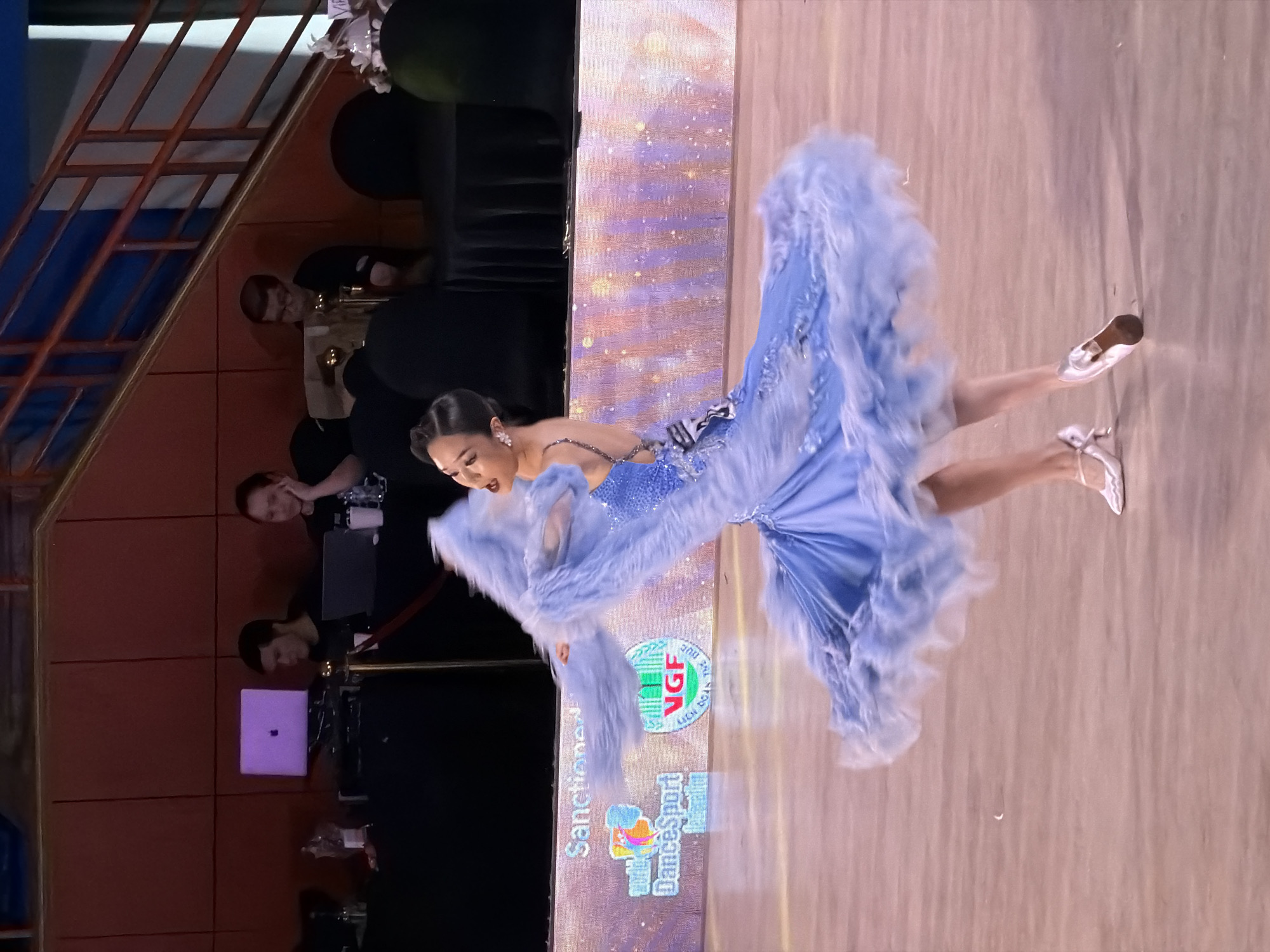 A female dancer in a flowing blue gown with feathery trim is captured mid-spin on a polished wooden dance floor. Her arms are extended, with one hand trailing a long, fluffy blue boa. The background features a digital screen displaying a logo that reads "World DanceSport Federation" and "VGF Lich Boa The Duc." Behind the screen, various individuals are seated at tables with laptops and equipment, suggesting an event or competition. The overall environment appears to be an indoor venue, possibly a convention center or auditorium, consistent with a dance competition setting. The lighting is bright, illuminating the dancer and the dance floor. This event is taking place at the National Convention Center in Hanoi, Vietnam.