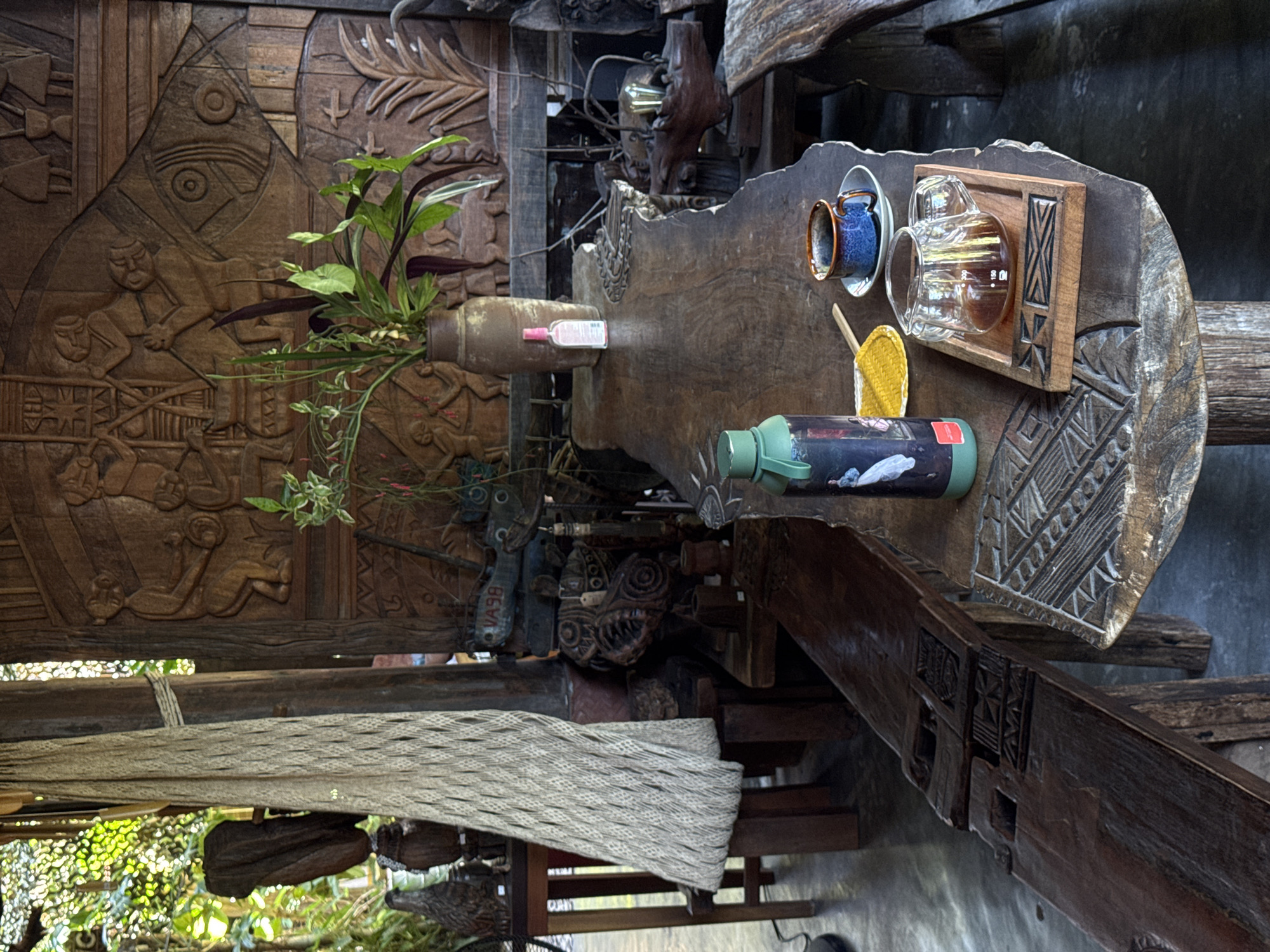 The image depicts a rustic wooden table setting in Đà Nẵng, Vietnam. The table is intricately carved with geometric patterns on its edge. Placed on the table are various items: a wooden tray holding a glass coffee server filled with coffee, a blue coffee cup on a saucer, a small yellow spatula, and a green water bottle with a design. A brown vase containing greenery stands next to the table on a wooden surface that also features carved figures. Below the table and vase, a woven fabric hangs from the wooden structure. In the background, there are additional carved wooden elements and glimpses of outdoor foliage, indicating a garden or courtyard setting.