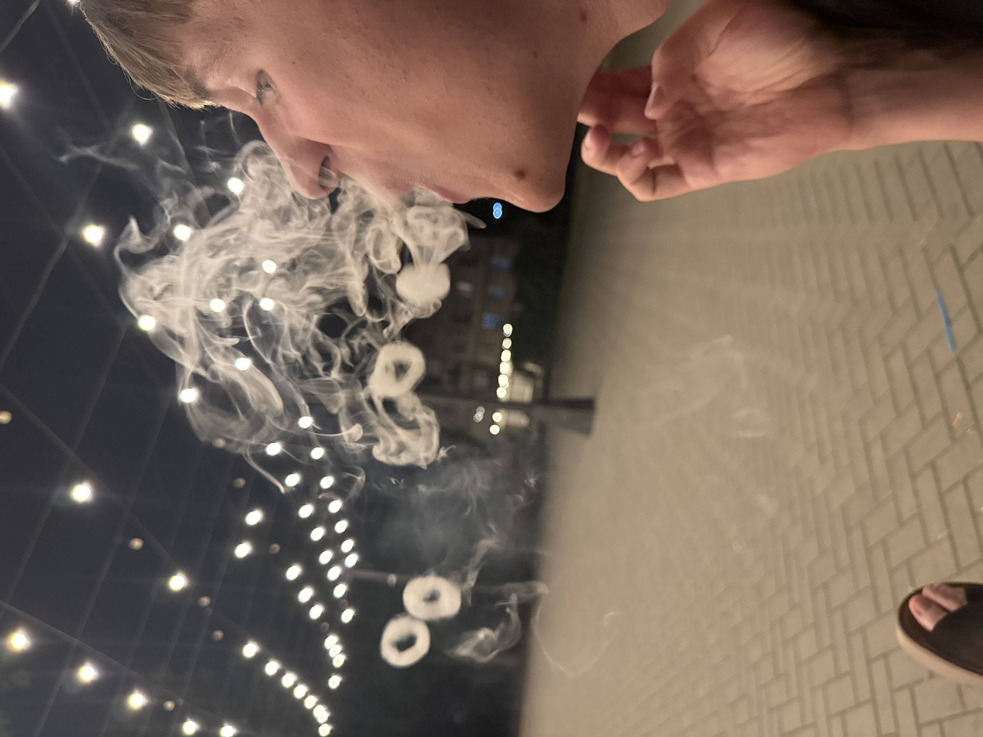 The image shows a close-up profile of a young person exhaling smoke, forming rings. The person's face is on the right side of the frame, with their mouth open. Smoke billows around their face and extends to the left, where several smoke rings are visible. In the background, there are strings of lights overhead, illuminating a paved outdoor area. The overall setting appears to be a public space at night, possibly a cafe or promenade. The details of the location in Tiraspol, Moldova, are not discernible from the image itself.