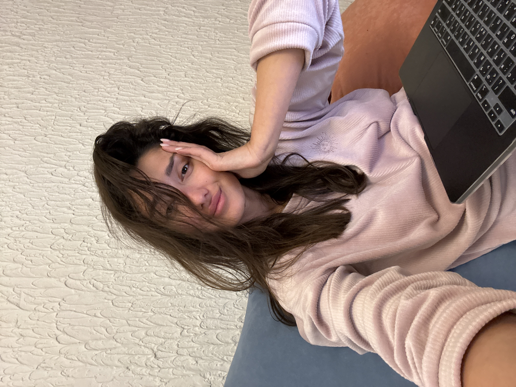 A woman is lying on her side on a blue couch, wearing a light pink sweater. She is holding a laptop in front of her and has her other hand on her forehead. The wall behind her is white with a textured pattern. The location of the image is unknown, the city of Sutomore, Montenegro cannot be determined from this picture.