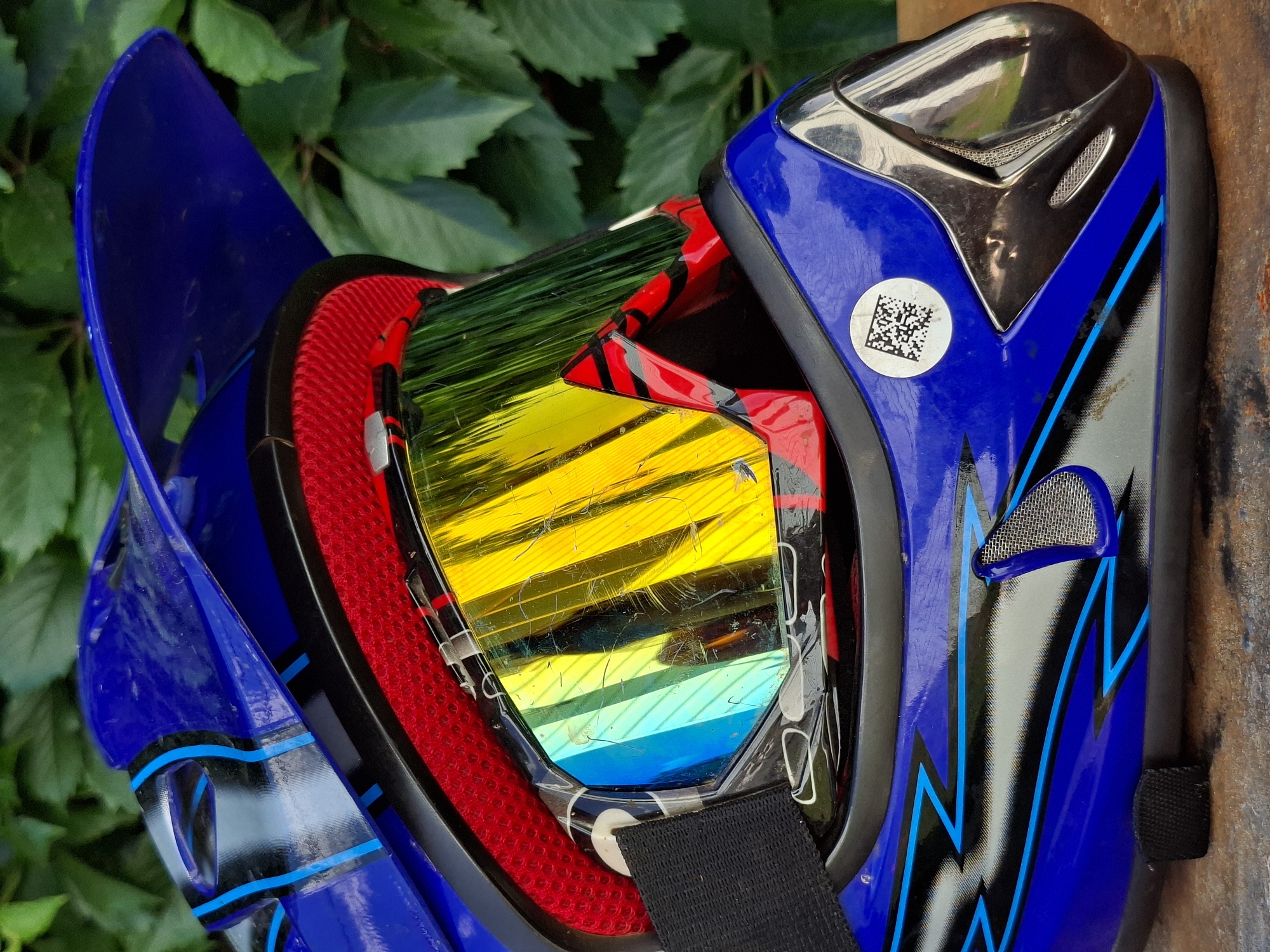 The image shows a close-up, side-view of a blue motocross helmet with red and black accents. The helmet features a prominent yellow and blue mirrored visor, which appears to have some scratches and scuff marks. A black goggle strap is visible attached to the side of the helmet. The helmet has a blue visor extending from the top and a black chin guard with mesh detailing. A circular sticker with a QR code is affixed to the side of the helmet. The helmet rests on a wooden surface, with a background of lush green foliage. The lighting suggests an outdoor setting, possibly in daylight. There are no people or animals present in the image. The location is unknown.