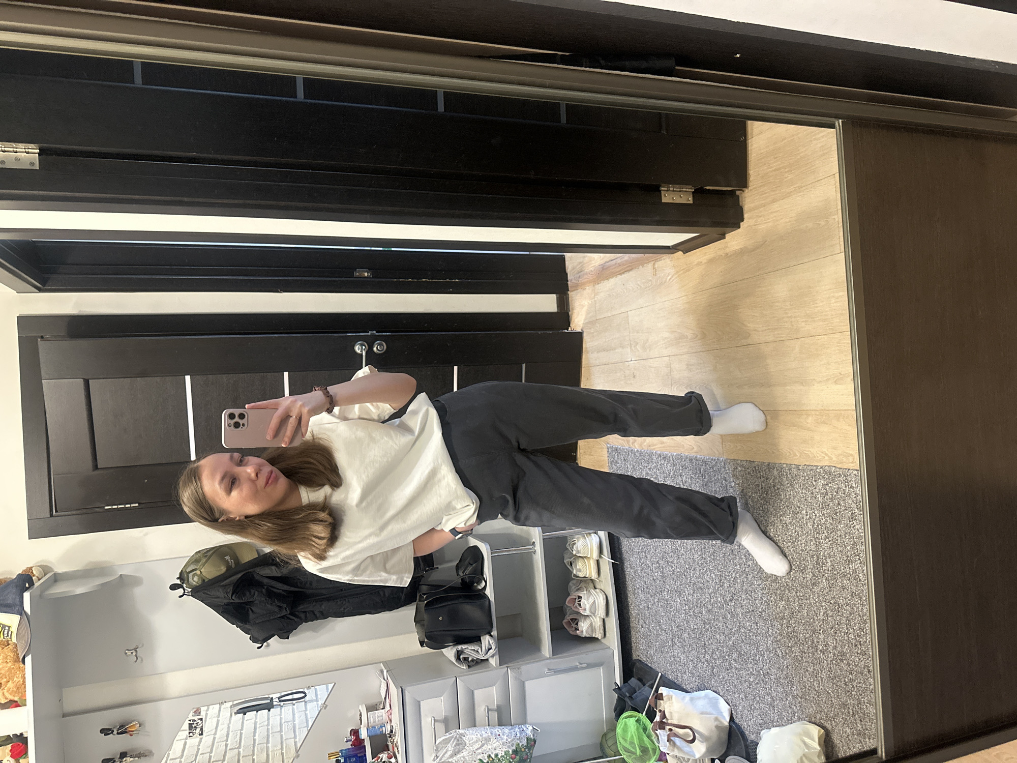 A young woman is seen taking a selfie in a mirror. She is wearing a white shirt, grey sweatpants, and white socks. She's holding a pink smartphone in her right hand. Behind her is a hallway with a dark wood-paneled door and a set of shelves containing shoes, bags, and other personal items. The floor is covered with a light-colored wood and a grey rug. Based on visual elements, the location could be in a residential apartment in Tomilino, Russia.