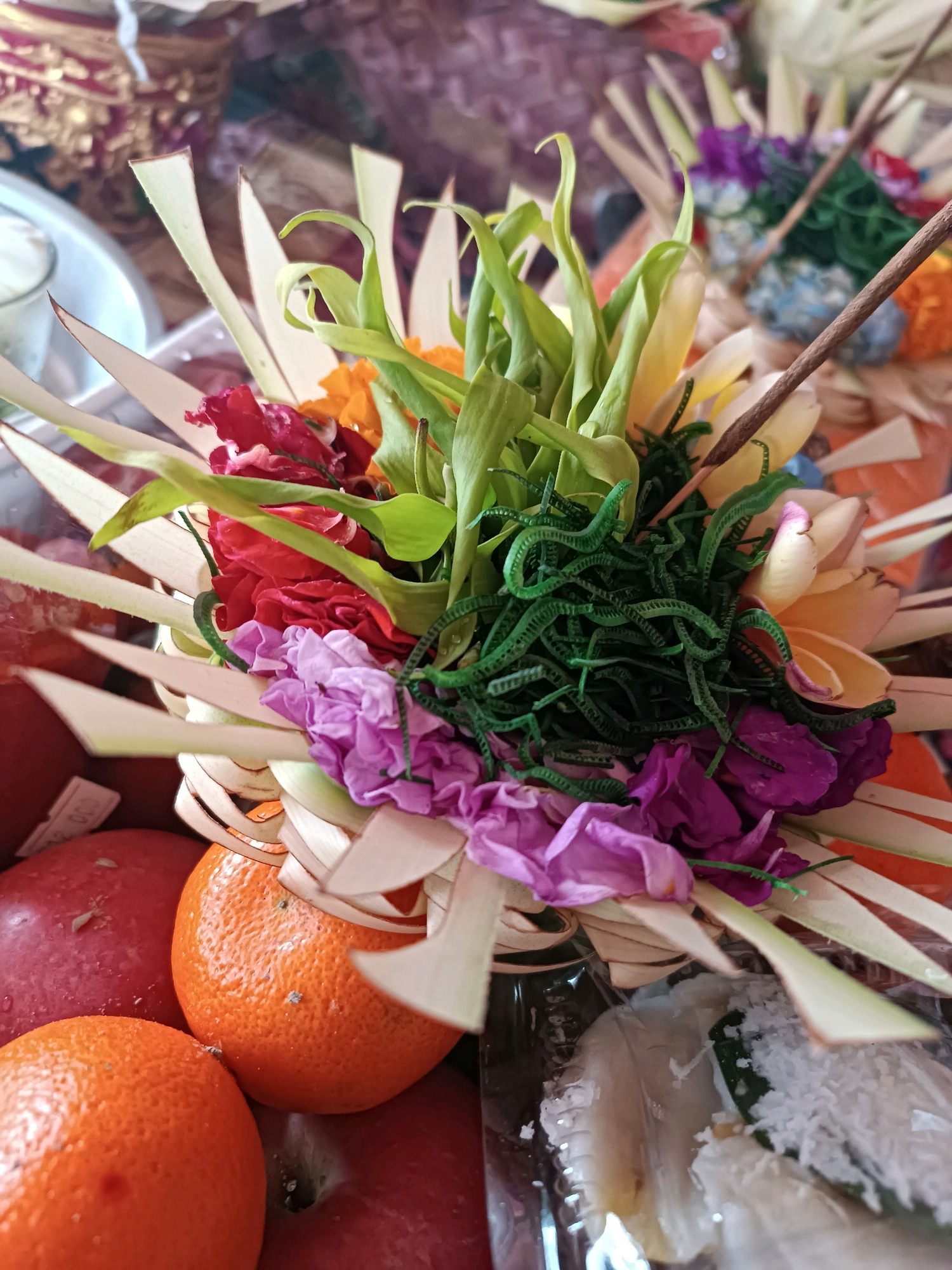 A close-up, vertical shot captures a decorative offering, likely a Canang Sari, featuring a vibrant arrangement of flowers and palm leaves. The foreground is dominated by red apples and oranges, with one orange nestled amongst the palm leaf elements of the offering. The offering itself is intricate, with tightly curled green fern-like fronds, delicate purple and pink flowers, and larger red blossoms. Yellow and light green floral elements are also visible. A thin, brown stick, possibly an incense stick, pierces through the arrangement. The background is blurred, showing more similar offerings and hints of a patterned textile. The scene appears to be set indoors, perhaps in a domestic or temple setting in Selemadeg, Indonesia, given the context of such offerings.