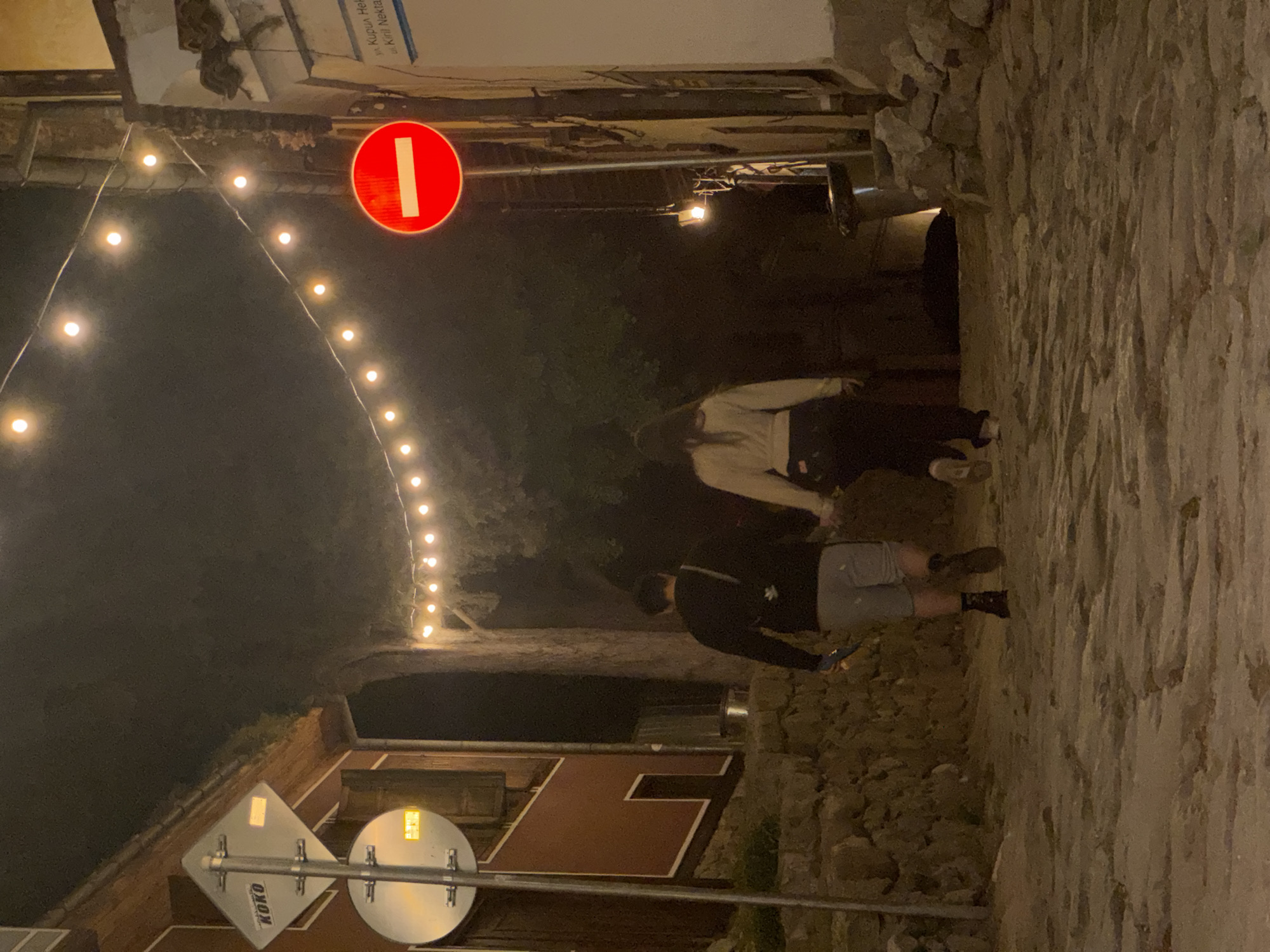 At night, the scene depicts a narrow alleyway in Plovdiv, Bulgaria, illuminated by a string of lights overhead. A red and white "no entry" sign is visible. Two individuals are present: one is wearing a light-colored top and dark pants, and the other is wearing a dark jacket and light-colored shorts. They appear to be walking through the alleyway. The path is lined with a stone wall on one side and buildings on the other. A sign with the word "KOKO" is also visible.