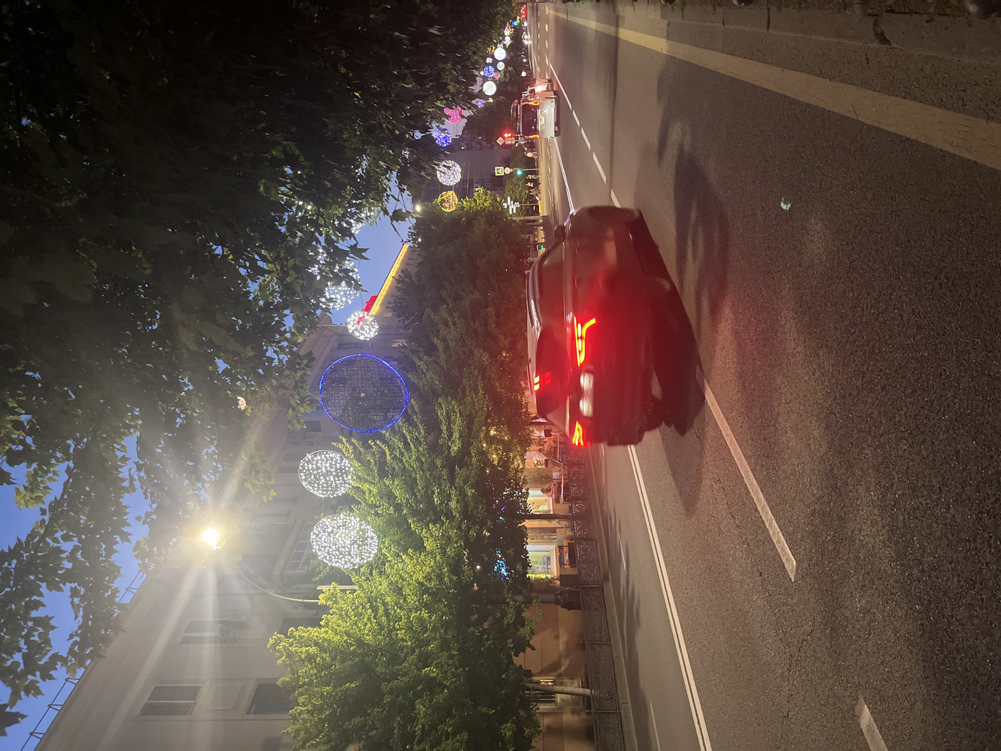 The image depicts a street scene at dusk in Krasnodar, Russia, likely during a festive season due to the elaborate string lights decorating the trees and buildings. A dark-colored SUV is in motion in the foreground, its taillights illuminated. The street is paved with asphalt and marked with white and yellow lines. On the left side of the street, there is a building with windows and trees. On the right side, more trees are visible, also adorned with lights. In the distance, other vehicles and streetlights can be seen, along with a few pedestrians on the sidewalk. The overall atmosphere is urban and lively, illuminated by artificial lights against the fading daylight sky.