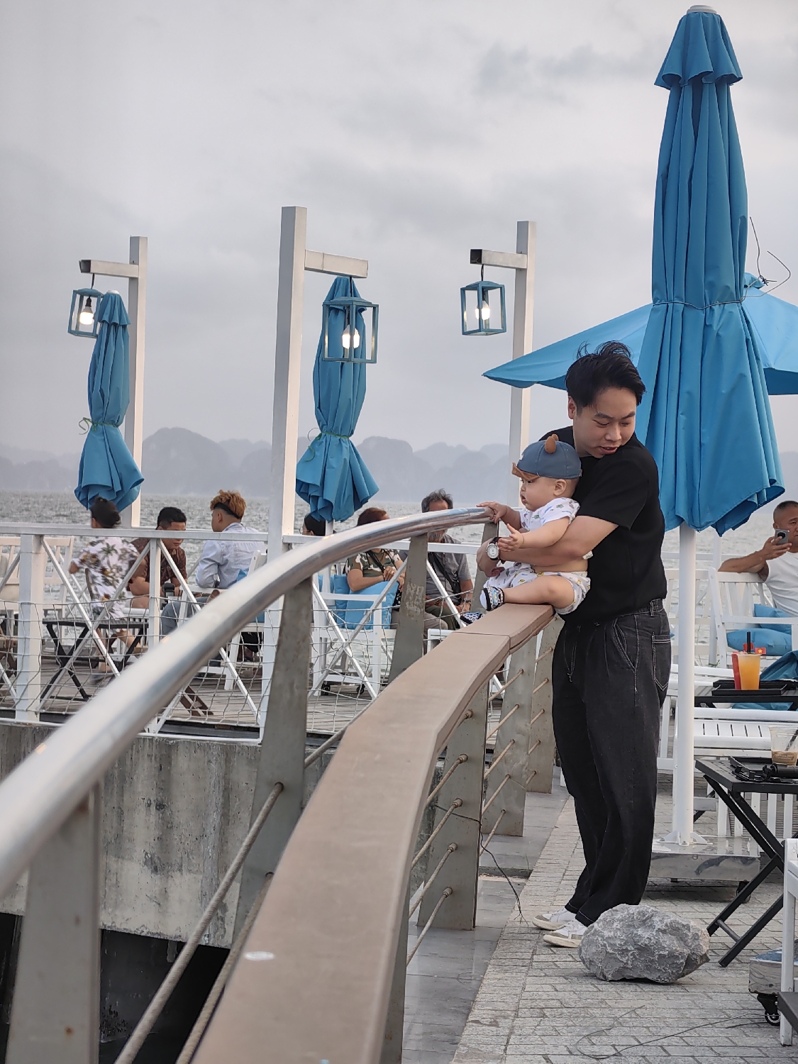 An Asian man holds a baby by a railing at an outdoor waterfront establishment. The man, wearing a black t-shirt and dark trousers, stands with the baby leaning over a curved brown and silver railing, looking out. The baby wears a light-colored onesie and a blue cap with bear ears.
In the background, several people are seated at tables, suggesting a cafe or restaurant setting. The area is equipped with white posts from which lit, lantern-style lamps hang, and bright blue umbrellas, mostly folded. Beyond the railing, a calm body of water is visible, with distinctive limestone karsts or mountains rising in the distance under an overcast sky. This view is characteristic of coastal areas like Bai Chay Ward in Vietnam, overlooking Ha Long Bay.
The soft, diffused light, combined with the illuminated lamps, indicates a late afternoon or early evening time. A large, grey, irregular rock rests on the patterned ground near the man's feet. The overall atmosphere appears relaxed and tranquil. No discernible text is visible within the image.