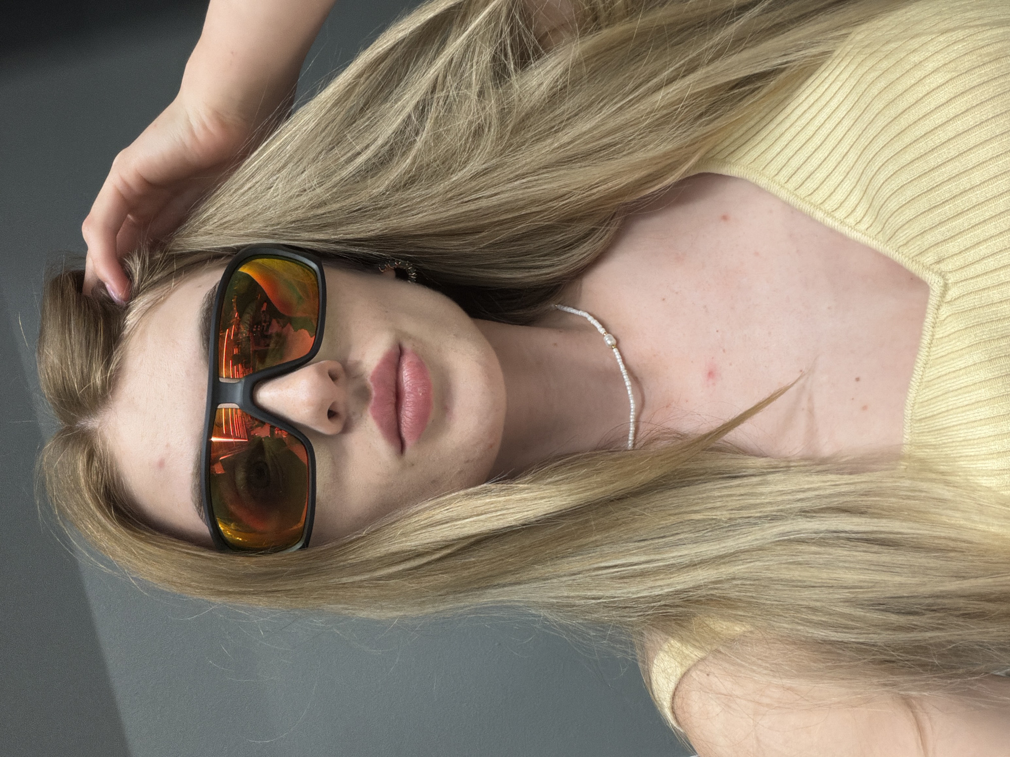 In an indoor setting with a plain gray wall as the backdrop, a young Caucasian woman with long, blonde hair is captured in a close-up, front-facing selfie. She is wearing large, black-framed sunglasses with orange-tinted lenses that reflect an interior environment with bright lights and some structural elements. Her right hand is raised, touching her hair near the top of her head. She has on a yellow, ribbed, v-neck top and a delicate white bead necklace. Her lips are slightly parted and painted with a light pink lipstick. The lighting suggests a dimly lit room, with the primary light source coming from above, casting subtle shadows.