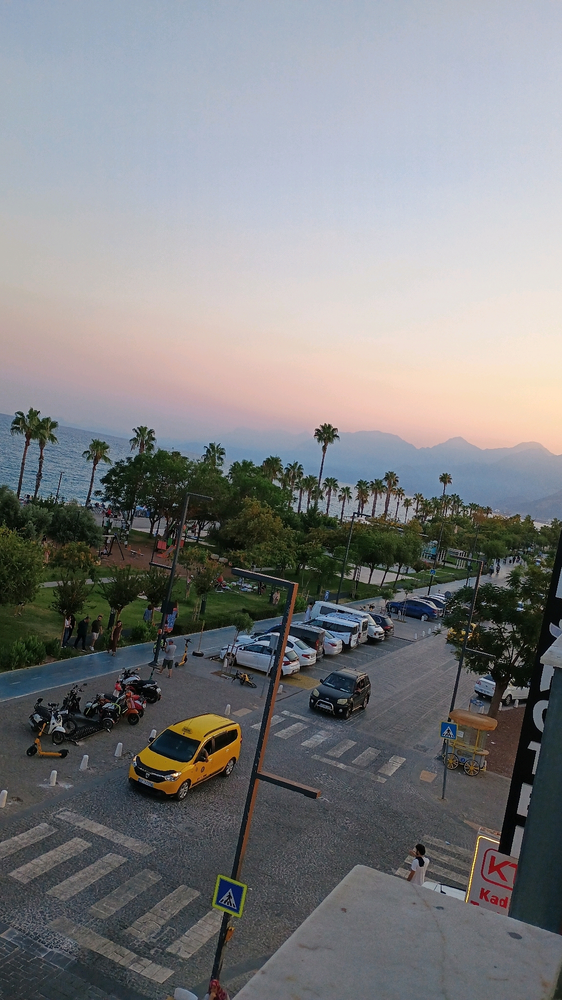 An aerial, high-angle, vertical shot captures a scene at sunset in Konyaaltı, Turkey. The sky transitions from a pale blue at the top to soft hues of pink and orange near the horizon, casting a warm glow over the landscape. In the background, the ocean stretches out, meeting a range of hazy, silhouetted mountains.
A prominent feature is a tree-lined promenade bordering the beach, with palm trees and lush green foliage. Several people are visible on the promenade, some walking, others sitting on the grass. Playground equipment is set up in one area.
The foreground displays a paved street with parked cars and motorcycles. A distinctive yellow taxi is in motion, traversing a pedestrian crossing marked with white stripes. Several other vehicles, including a black SUV, a white van, and various cars, are parked along the roadside. Three scooters are parked near some motorcycles. Pedestrian crossings and traffic signs are visible on the street.
The image also shows the edge of a building in the lower right corner, with a glimpse of signage. The overall atmosphere is serene, with the soft lighting of sunset enhancing the coastal and urban elements of the scene.
