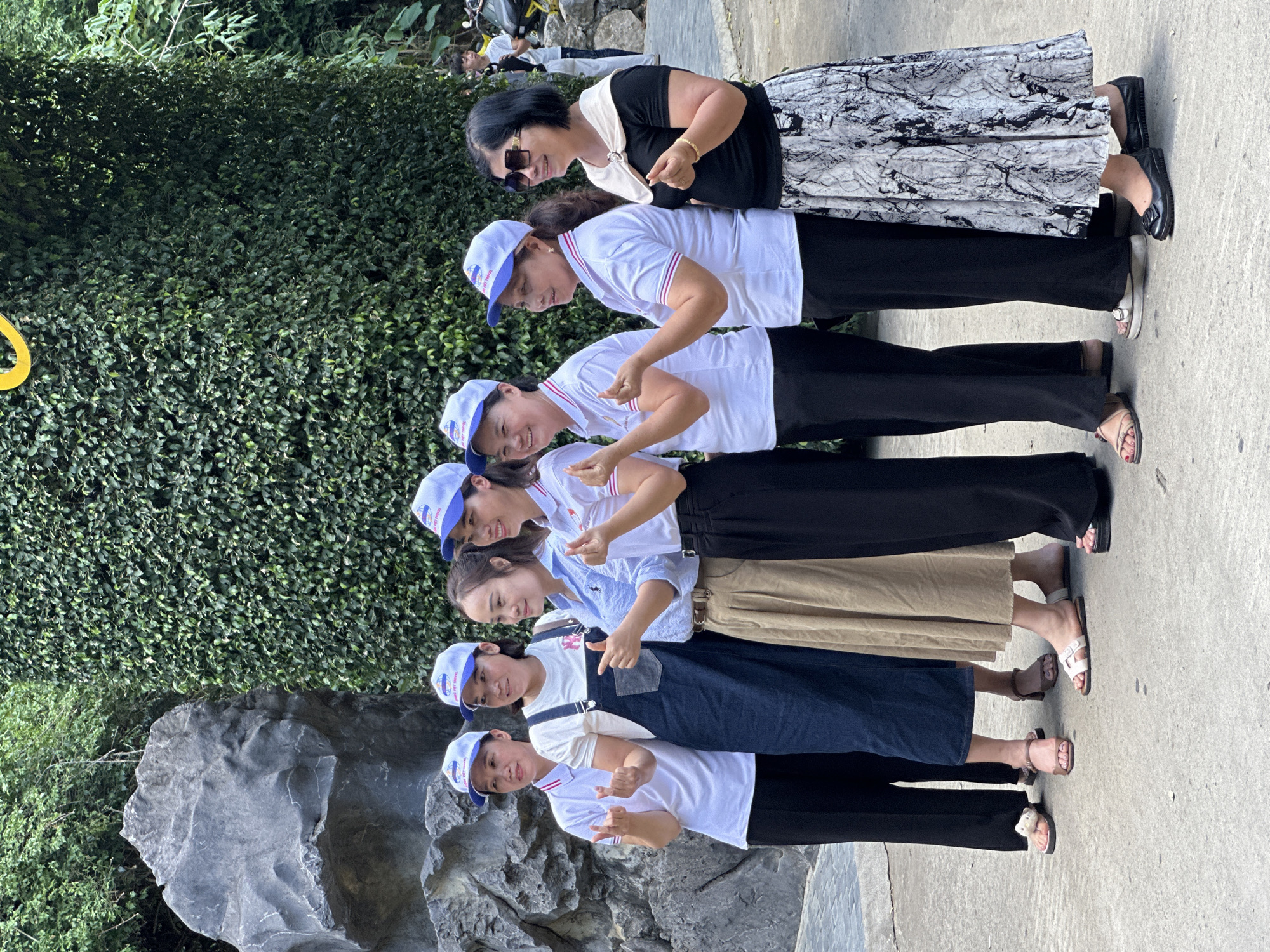 A group of seven women, all wearing matching blue and white baseball caps and white polo shirts, stand in front of a tall, dense green hedge wall and a large grey rock formation. The women are posing for a photograph, with several of them making heart shapes with their fingers. The woman on the far right is wearing a black top and a black and white patterned skirt, and sunglasses. The woman second from the left is wearing a dark blue denim overall dress over her white polo. The woman third from the left is wearing a tan skirt. The ground appears to be a paved walkway. The environment suggests an outdoor tourist location, possibly a garden or park. The woman on the far right appears to be the oldest in the group. The woman in the denim overalls appears to be the youngest. The group is likely posing for a photograph while on a tour of the city of Yên Sơn Ward, Vietnam.