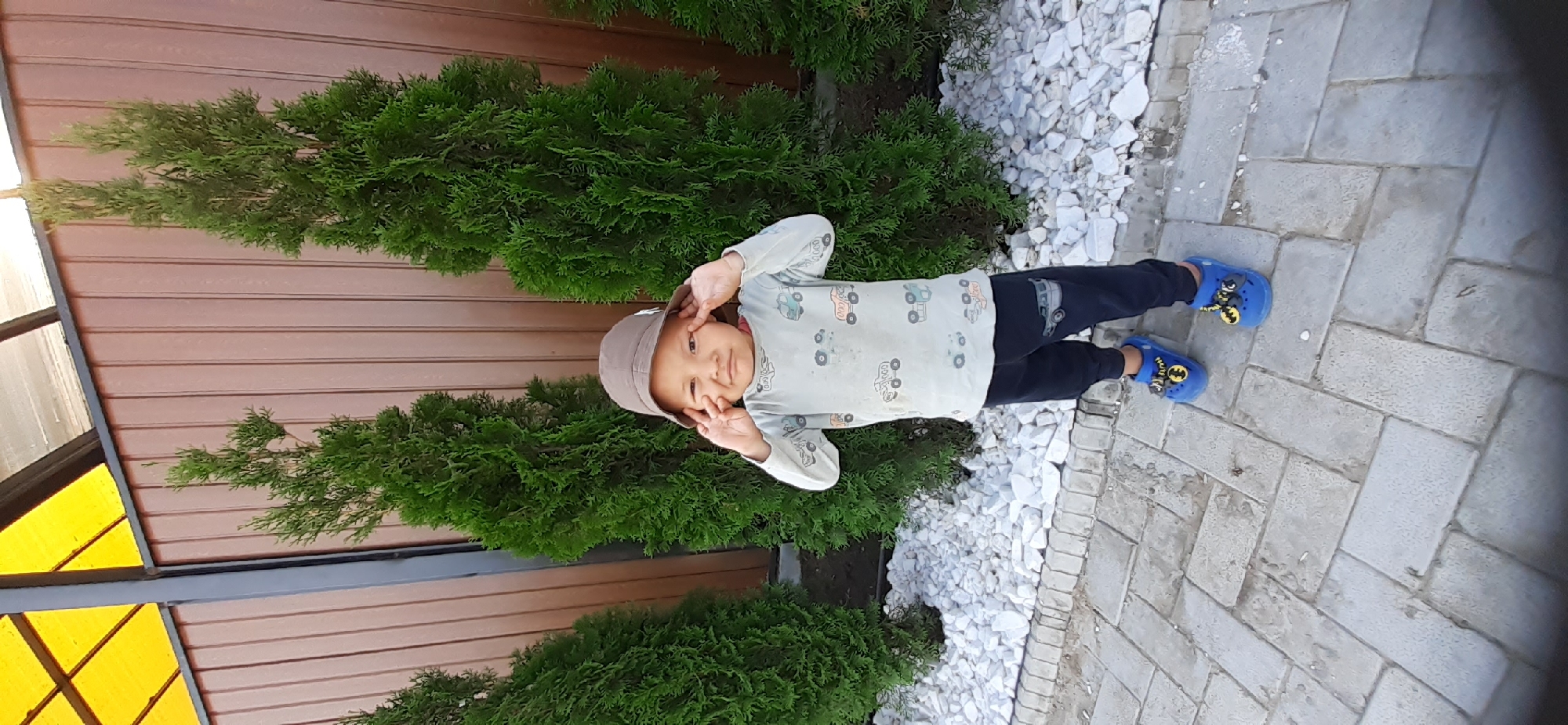 A young child wearing a brown hat, a light-colored shirt with a car print, and dark pants stands outside in front of a brown wooden fence. The child is holding their hands up to their face, with fingers extended near their eyes, making a playful gesture. Behind the child are several tall, green evergreen trees. The ground in front of the fence is covered with white pebbles, and further forward, a paved pathway made of gray interlocking stones is visible. A yellow awning can be seen above the fence. The scene appears to be in a garden or outdoor area, possibly in Choktal, Kyrgyzstan.