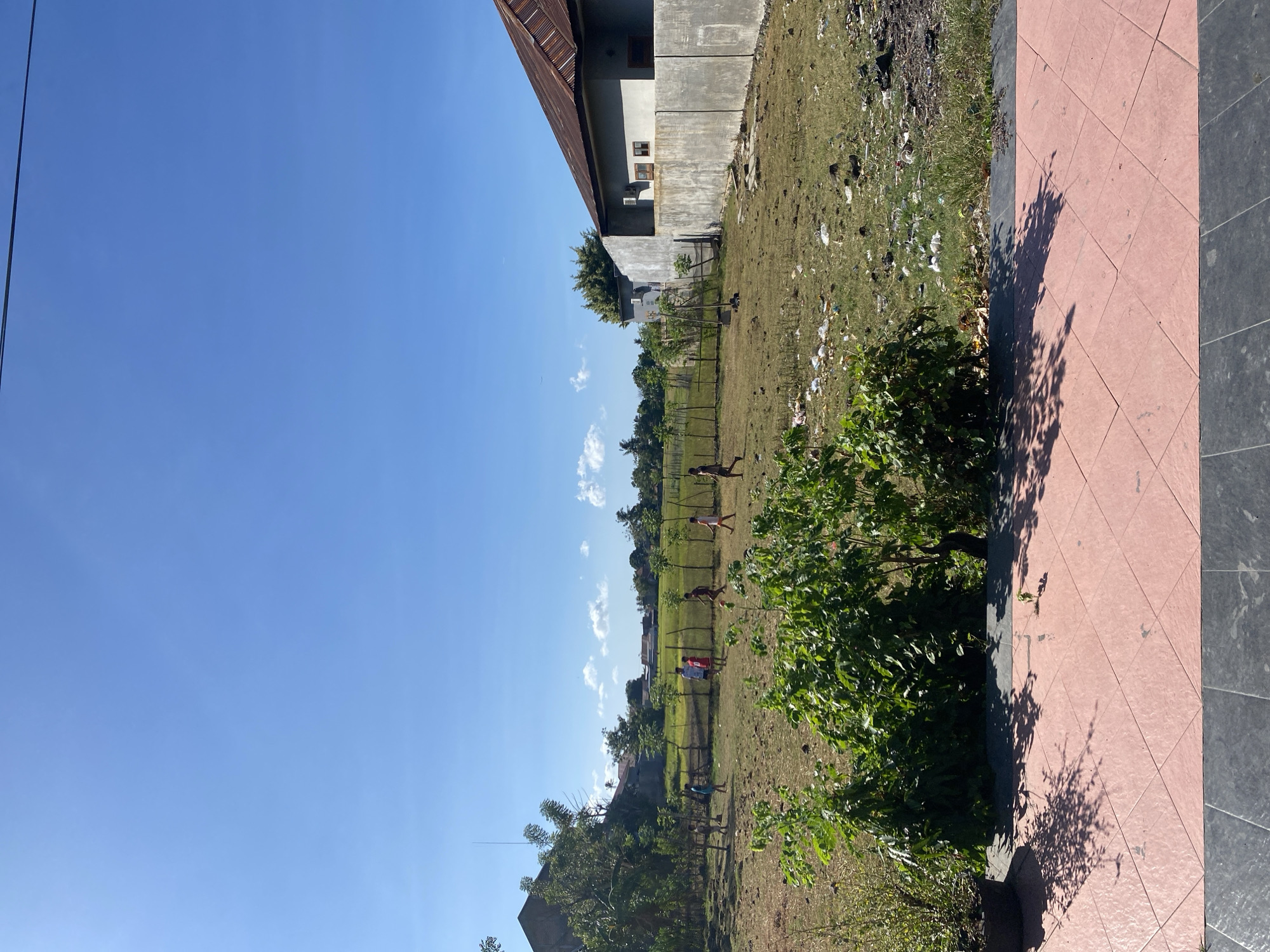 The image depicts a sunny day in a residential area of Bantaeng, Indonesia. A group of children are playing soccer in a dry, grassy field. In the background, there are houses with traditional Indonesian architecture, some with corrugated metal roofs. A fence separates the field from a neighboring property, which appears to be undergoing construction or renovation. The ground in the foreground is paved with pink and gray tiles, and lush green bushes are in the foreground. The sky is clear blue with a few scattered clouds. The overall scene suggests a typical afternoon in a rural Indonesian village, where children find joy in simple outdoor activities.