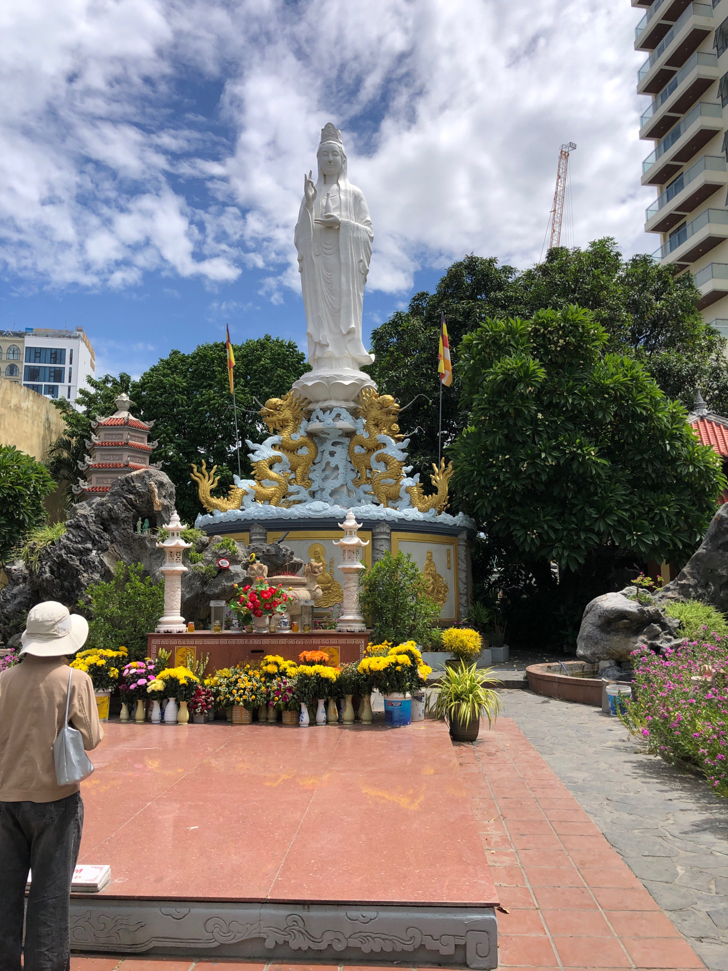 The image captures a daytime scene at a Buddhist temple in Da Nang, Vietnam. A large, white statue of a deity, possibly Quan Am, stands prominently in the center, adorned with golden dragons and clouds at its base. In the foreground, a person wearing a wide-brimmed hat and a brown shirt faces the statue, their back to the camera. They appear to be observing or praying.
Surrounding the central statue are various elements typical of a Vietnamese temple: colorful flowers, incense burners, and smaller statues or decorative elements. A tiered pagoda structure is visible to the left, and a modern apartment building and a construction crane rise in the background, suggesting the temple is located in an urban or developing area. The sky is blue with scattered white clouds. The ground is paved with red tiles in the immediate foreground, transitioning to a stone pathway on the right, lined with more greenery and rocks. The overall atmosphere suggests a place of worship and contemplation within a bustling city.