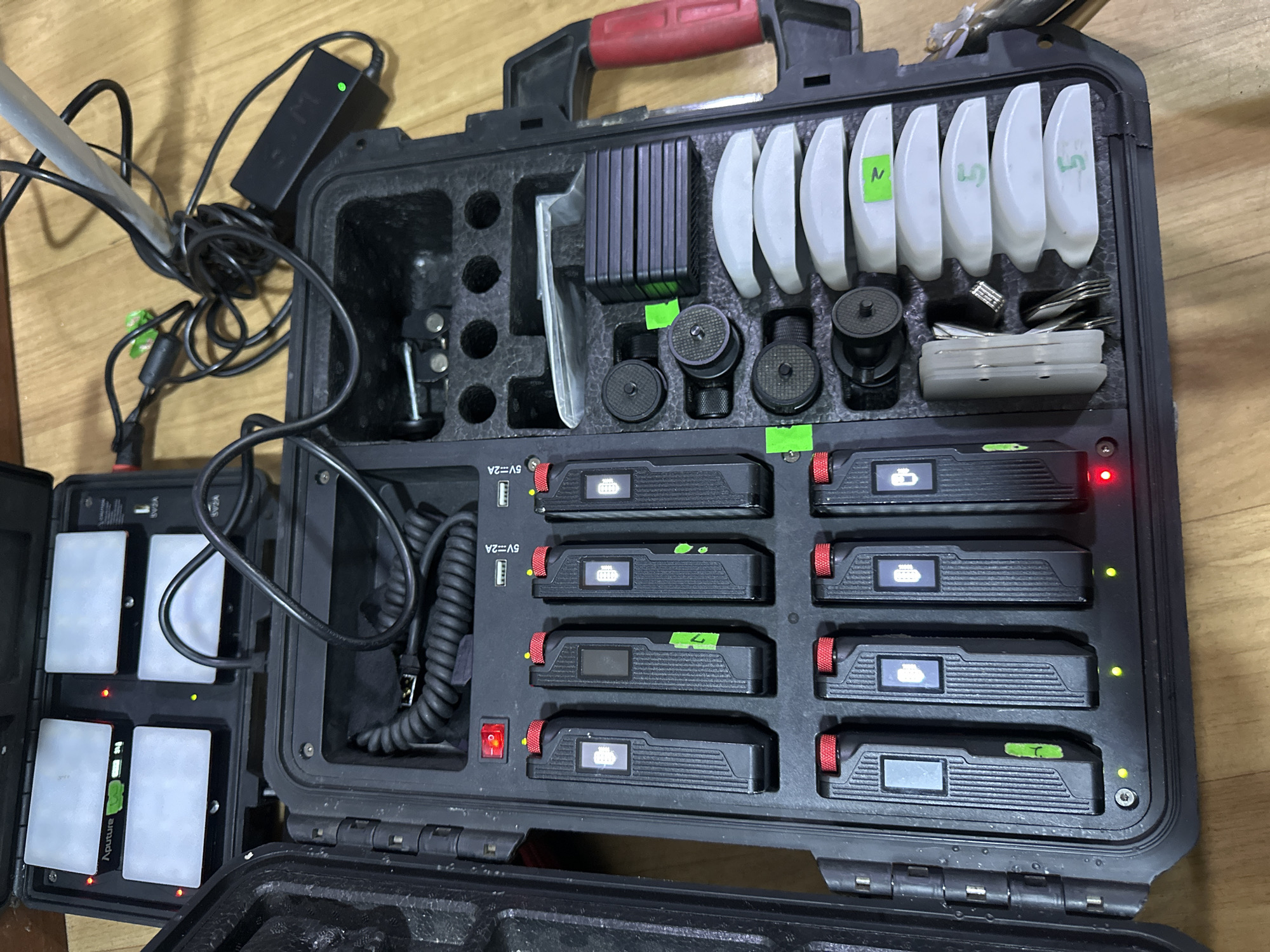 A black equipment case is open, displaying various electronic components. The case is divided into compartments containing items such as batteries, chargers, cables, and small accessories. Some of the batteries are labeled with numbers. Power adapters and cables are visible, suggesting a charging or operational setup. The environment is a wooden surface, likely a floor or table. There is no indication of the location being in Lagos, Nigeria. The description focuses on the visible contents and lacks the elements necessary to assert the location or include individuals.