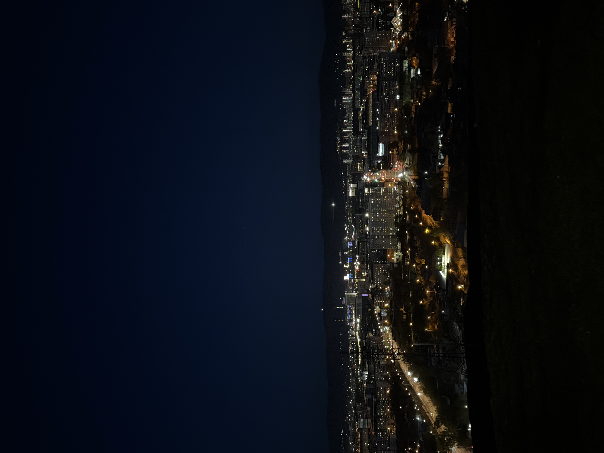 A nighttime cityscape is captured from an elevated position, likely a hilltop or high-rise building, providing a panoramic view of Krasnoyarsk, Russia. The city lights illuminate the urban sprawl, revealing a network of roads and buildings. The sky is dark, contrasting with the warm glow of the city below. In the distance, a silhouette of a mountain range is faintly visible. The foreground has a dark area to the right, suggesting a closer element such as vegetation or a wall, from the viewing location.