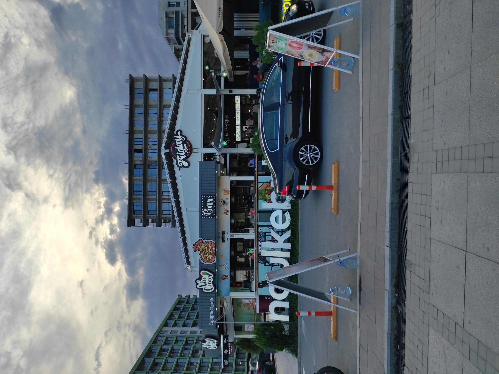 The image depicts a street scene in Năvodari, Romania, featuring a commercial establishment with outdoor seating. In the foreground, a black sedan is parked on the asphalt. Two A-frame signs with informational content are positioned in front of the vehicle. To the left, large white letters spell out "ngulkeber". Behind the signs and car, a row of businesses is visible, including "Vice Cream", a "Bar", and "Friday Pizza". The architecture of the buildings varies, with some appearing to be under construction or recently built, characterized by modern apartment complexes and a multi-story building with unfinished facades. The sky is overcast with dramatic clouds. People are seated at tables outside the establishments, suggesting a public space with dining and recreational activities. The overall environment appears to be a coastal or waterfront area, common in resort towns like Năvodari.