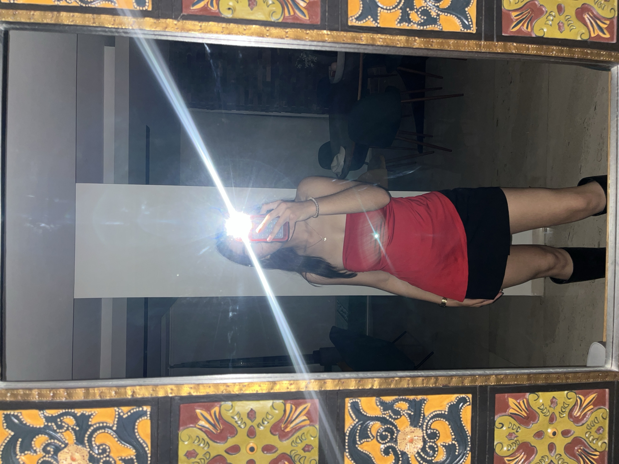 An indoor mirror selfie captures a person rotated 90 degrees, appearing horizontally in the frame. The subject, seemingly a young woman, is wearing a red strapless top, a black mini skirt or shorts, and black knee-high boots. She holds a smartphone, taking the picture, with a bright flash obscuring her face and creating a strong glare across the mirror. Her left wrist displays a thin bracelet, and she wears a delicate necklace.
The mirror itself is encased in a striking, ornate frame. This frame features a metallic gold border and square tiles adorned with intricate, colorful patterns in yellow, black, red, and blue.
The background reflection, largely obscured by the person and the flash, shows hints of a dimly lit interior. Elements resembling dark furniture, possibly chairs and tables, are visible, along with light-colored walls or architectural features. The overall setting is an indoor space, likely a home or establishment, possibly in San Bernardino Tlaxcalancingo, Mexico, given the traditional tile design of the mirror frame. No visible text is present in the image.
