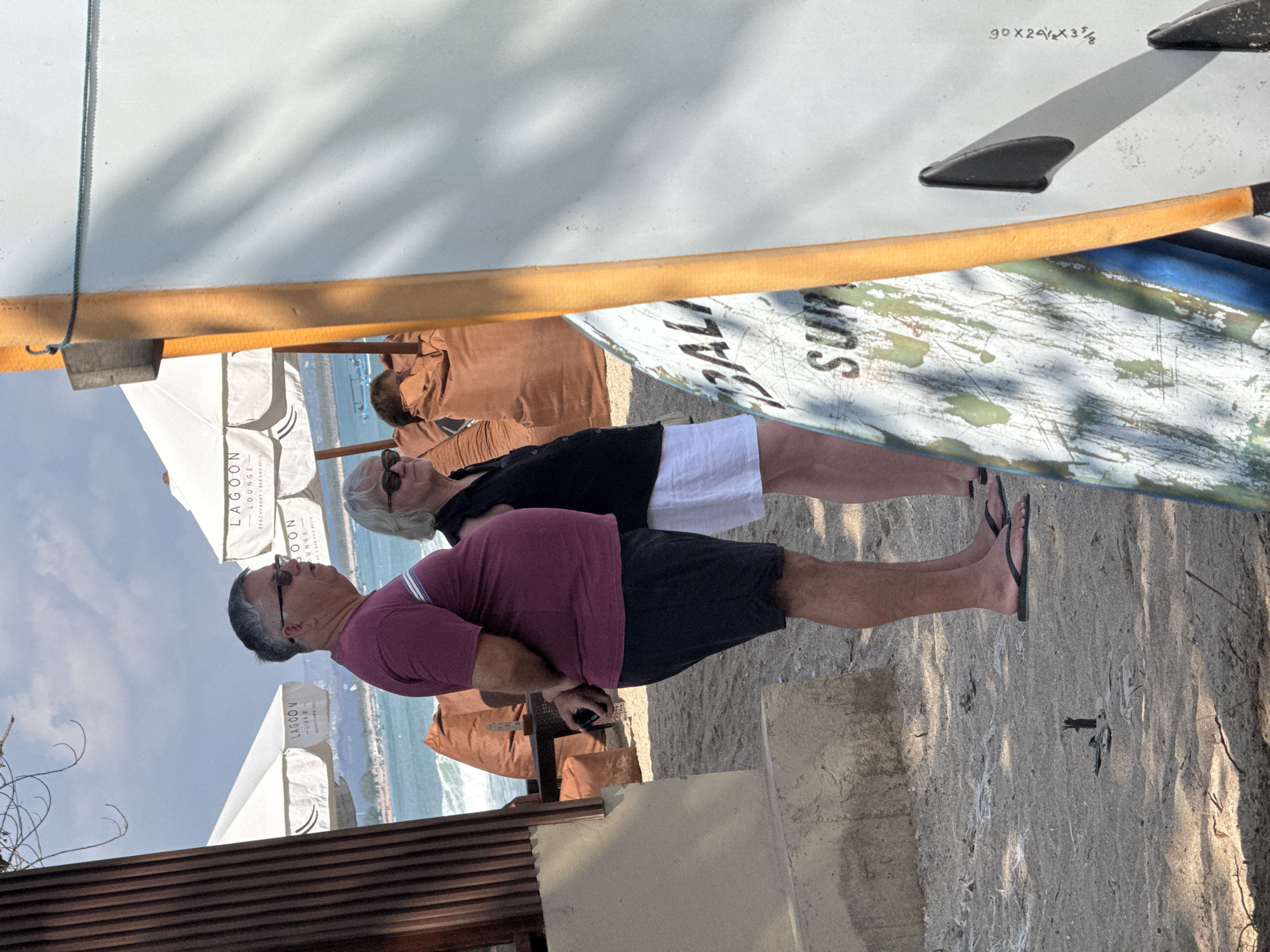 In Kuta, Indonesia, on a sandy beach, a man and a woman stand near umbrellas and beanbag chairs. The man, wearing a maroon t-shirt and dark shorts, has his hands behind his back. The woman, in a black top and white skirt, stands beside him. A surfboard leans against a wooden pole in the foreground, obscuring part of the scene. The ocean and waves are visible in the background under a cloudy sky.