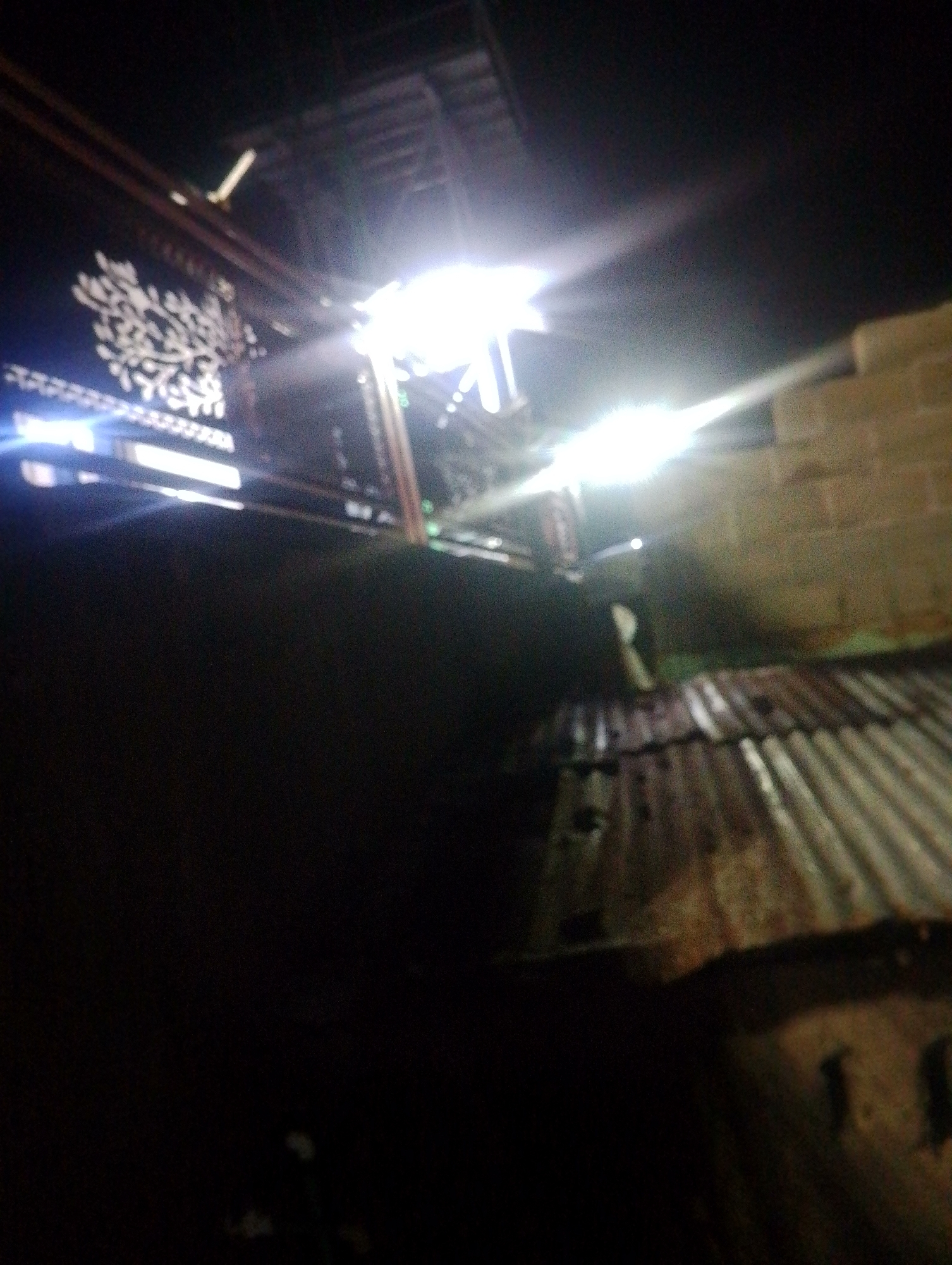 The image depicts a dark, outdoor scene, likely at night, with several sources of bright light causing lens flare and overexposure. In the foreground, a dark, possibly textured surface dominates the lower half of the frame. Above this, a corrugated metal roof is visible, showing signs of wear and rust. To the left, a dark structure with a white, intricate pattern, possibly a decorative screen or facade, is illuminated by bright lights. These lights emanate from an unseen source above and to the right, casting strong beams that streak across the image. In the background, a brick wall can be seen. The overall impression is of a dimly lit urban environment at night, possibly a residential or commercial area. Without further context or clearer details, pinpointing the exact location within Jos, Nigeria, is not possible. The scene appears to be an unadorned, utilitarian setting, characteristic of many parts of the city.
